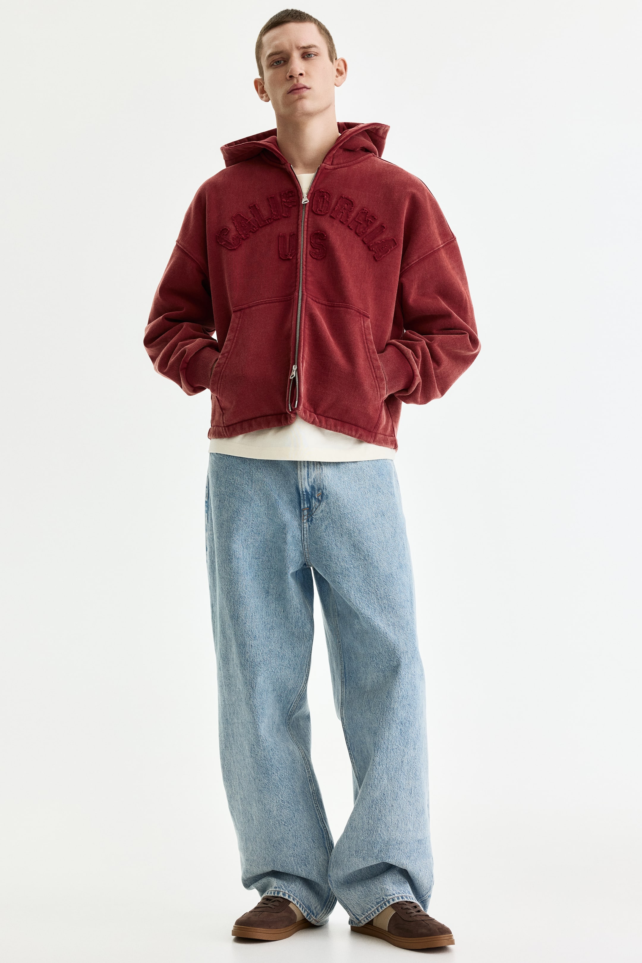 View larger image: A washed red zip-up hoodie features a full-zip closure down the front and "CALIFORNIA US" in raised embroidered lettering on the chest. It also has front kangaroo pockets, ribbed cuffs, and a ribbed hem, and is worn partially unzipped over a white T-shirt.