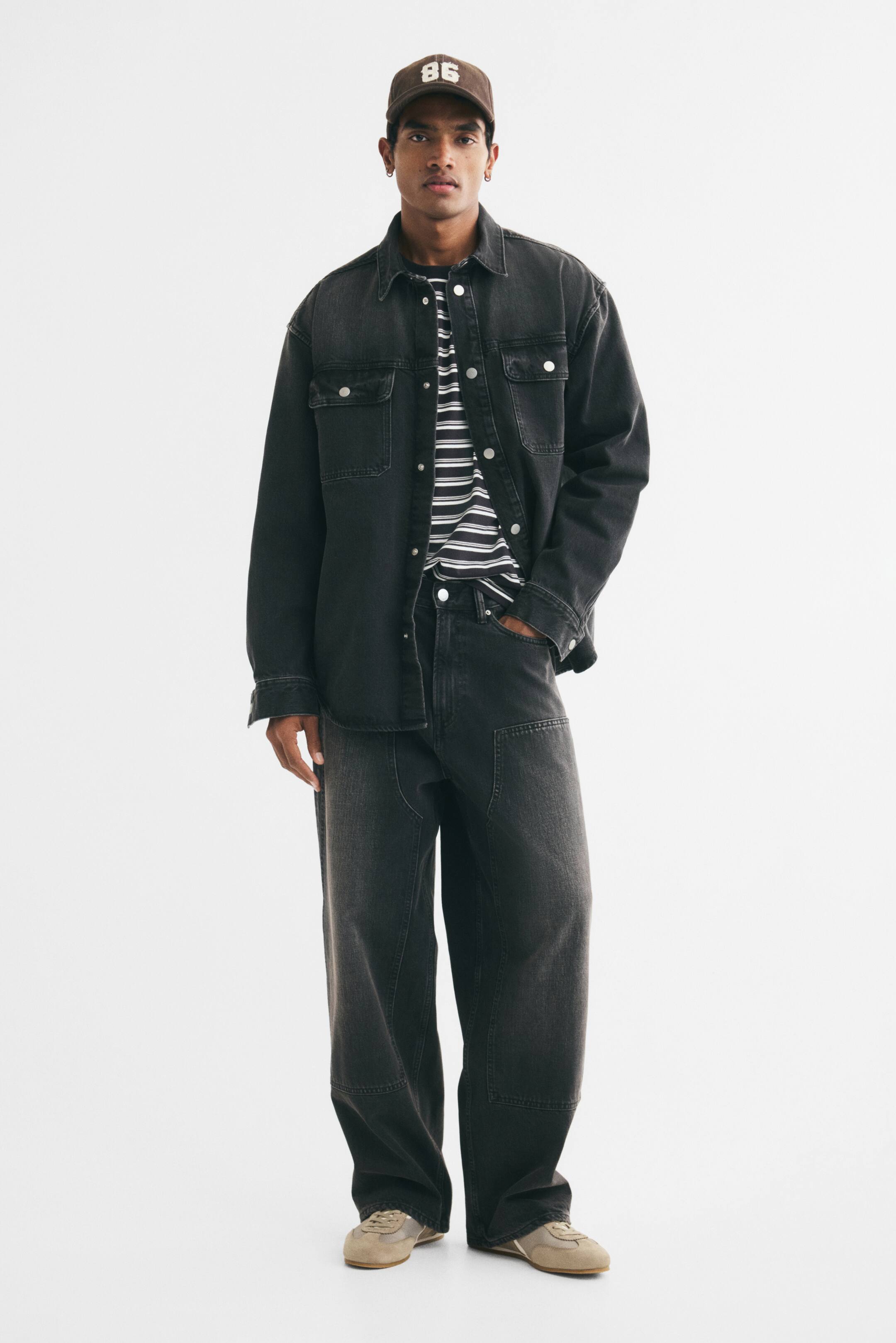 View larger image: The model is wearing an outfit featuring a black denim overshirt, styled open over a black and white striped T-shirt. The overshirt has a pointed collar, button front, and two chest pockets, paired with black denim pants with knee panel details, a brown baseball cap, and light-coloured sneakers.
