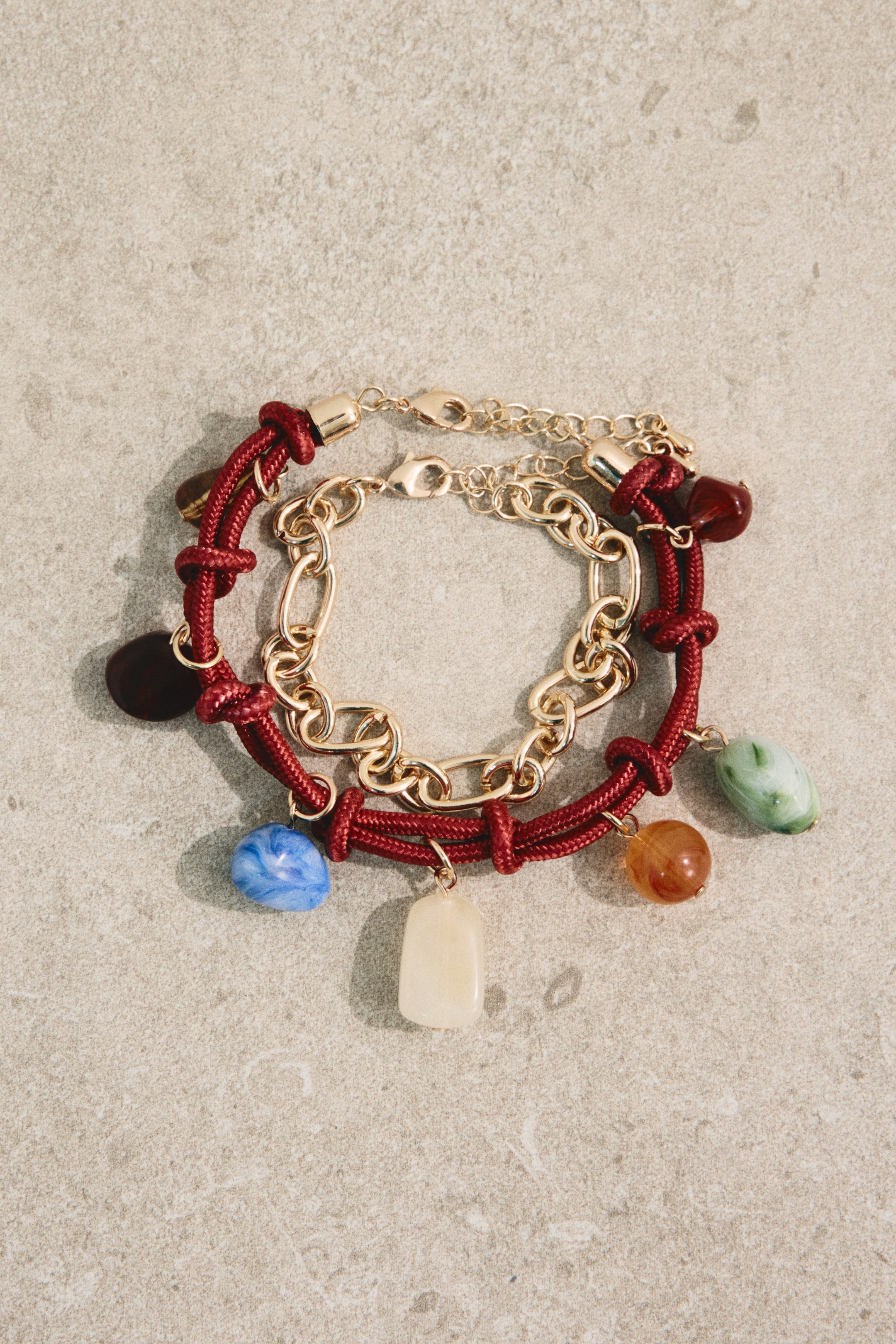 View larger image: Two bracelets are positioned at an angle. One is a red cord bracelet showcasing multiple colorful, irregularly shaped charms and a gold-tone lobster clasp. The other is a gold-tone chain link bracelet with large oval links, also featuring a gold-tone lobster clasp.