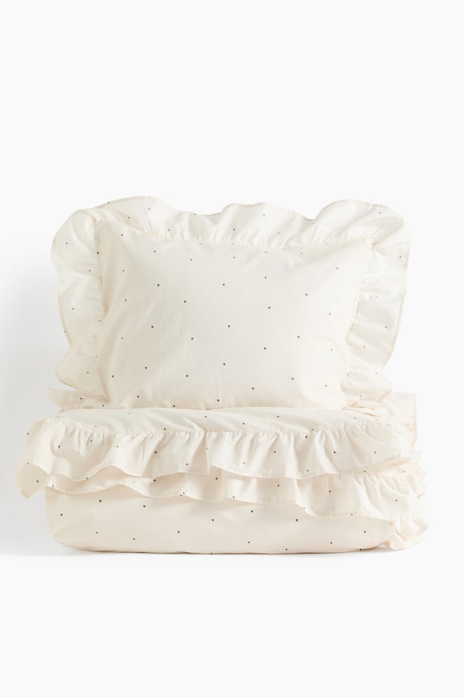 Een crÃ¨mekleurige beddenset, met een kussensloop en een opgevouwen dekbedovertrek aan de voorkant, met een patroon van kleine, verspreide donkere hartjes. Beide stukken hebben opvallende rucheranden die een zachte, decoratieve textuur toevoegen. - 1