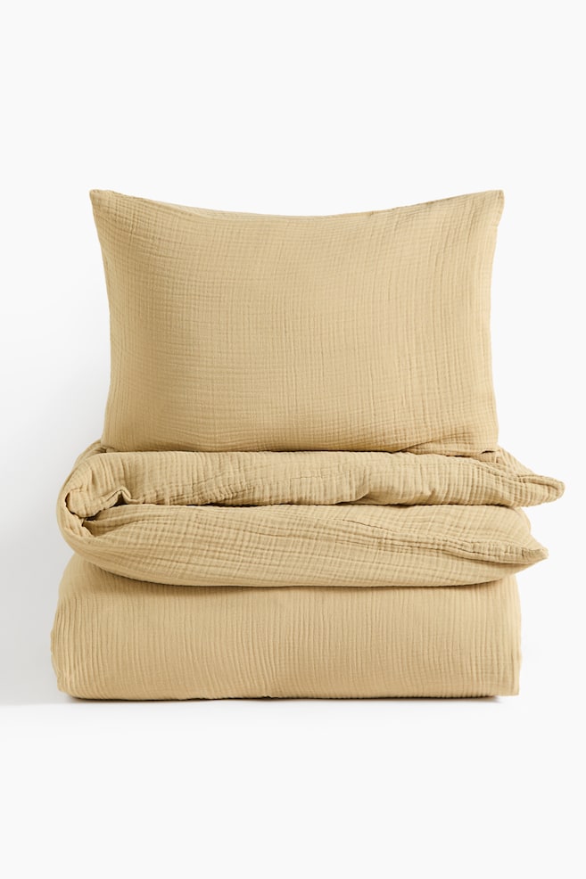 A single duvet cover set in warm beige cotton muslin is facing forward, showcasing its crinkled texture. The matching pillowcase is resting upright on top of the folded duvet cover. - 1