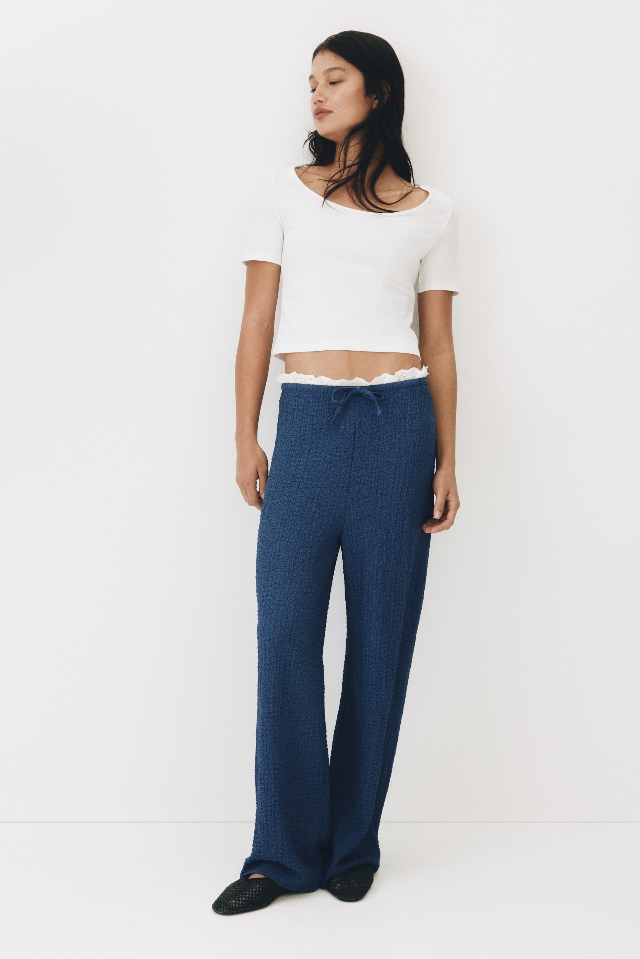 Xem ảnh lớn hơn: Quần ống rộng bằng vải jersey xanh nhăn có cạp chun, dây rút với viền bèo nhỏ được trang trí bằng ren broderie anglaise. Người mẫu đang mặc chiếc quần này.