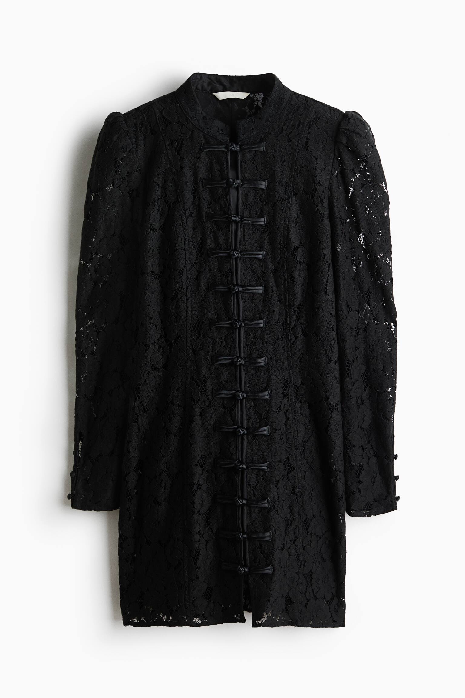 A short, fitted black lace dress is shown from the front, featuring a band collar and a vertical row of satin-style knot buttons down the centre. The dress features long puff sleeves with small covered buttons at the cuffs and visible vertical seams running down the front of the bodice.