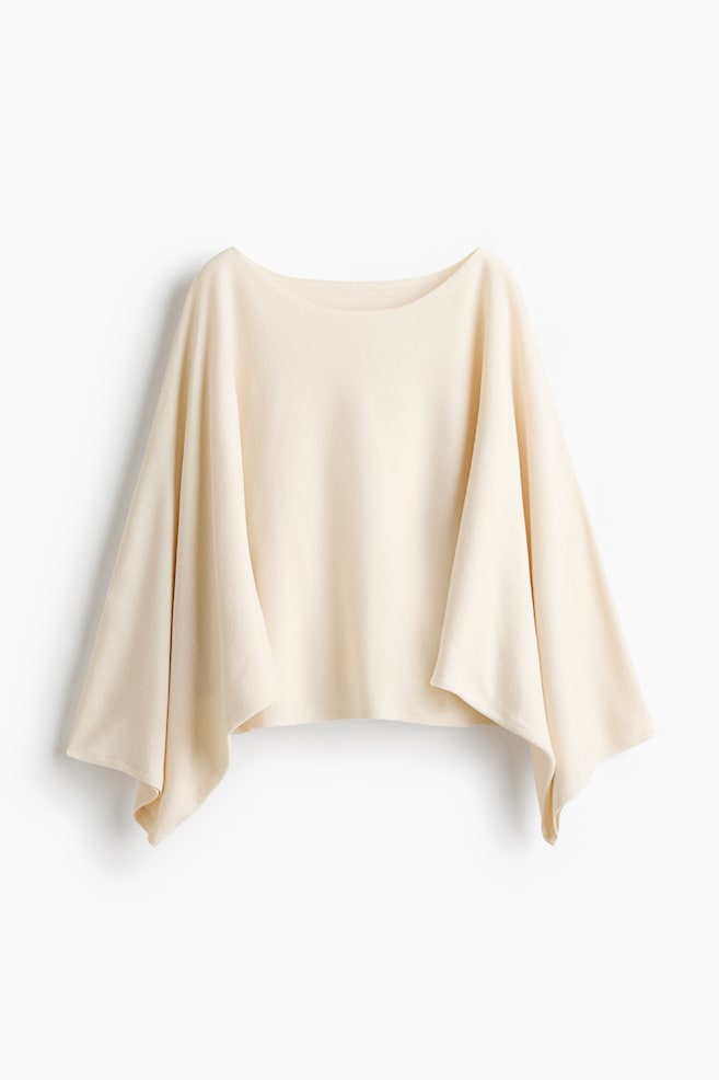 A cream-colored, short, loose-fit knit top is shown in a front-facing view, featuring a delicate boat neckline and long, draped cape sleeves. The soft, fine-knit fabric is flowing loosely on both sides, emphasizing its relaxed and airy silhouette. - 1