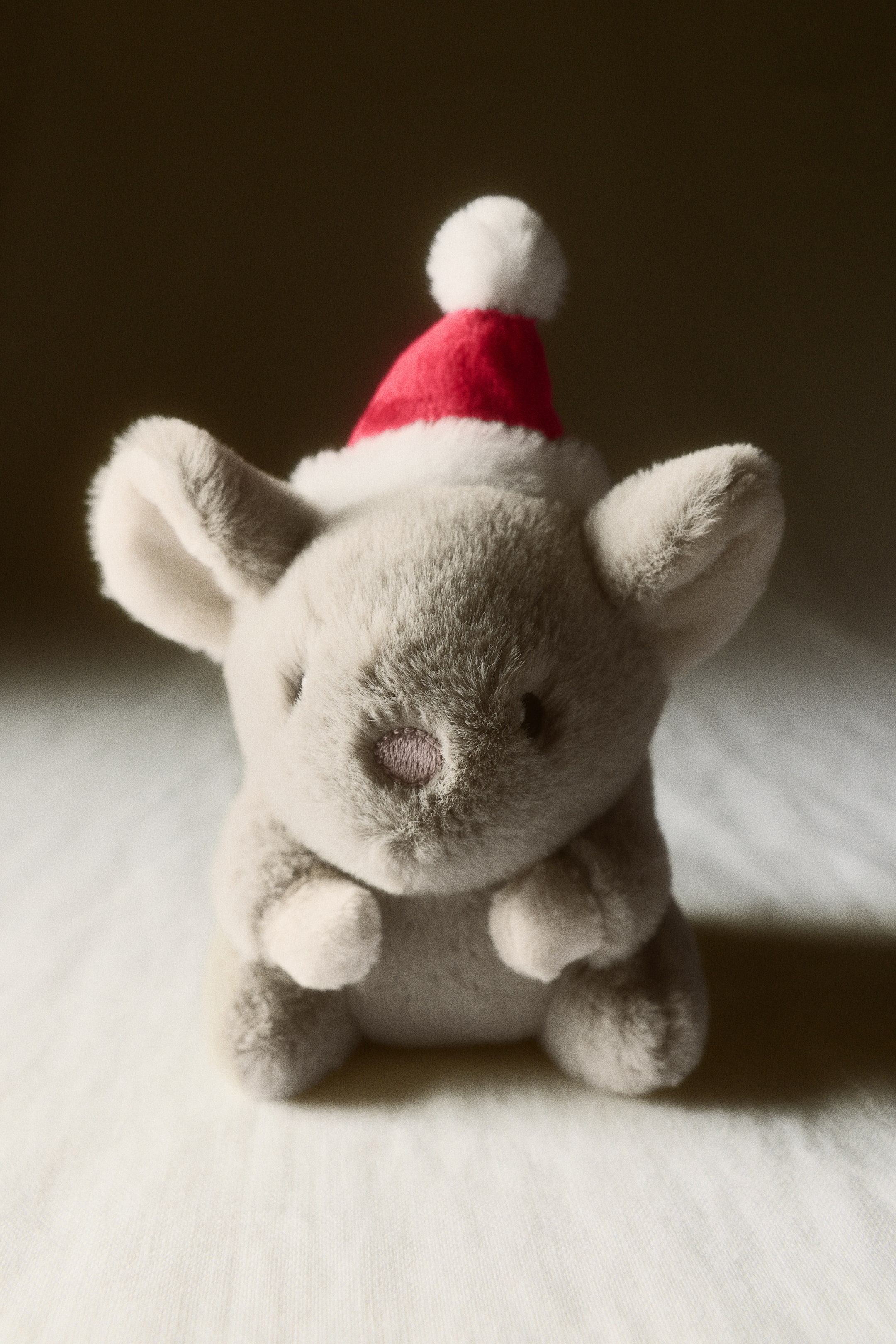 Grösseres Bild ansehen: Ein weiches, weihnachtlich gestaltetes Plüschtier mit einem hellgrauen Plüschtier-Tier sitzt und blickt nach vorne. Das Spielzeug trägt eine rote Weihnachtsmütze mit weissem Besatz und Bommel und hat kleine, aufgestickte schwarze Augen sowie eine pinke Nase.