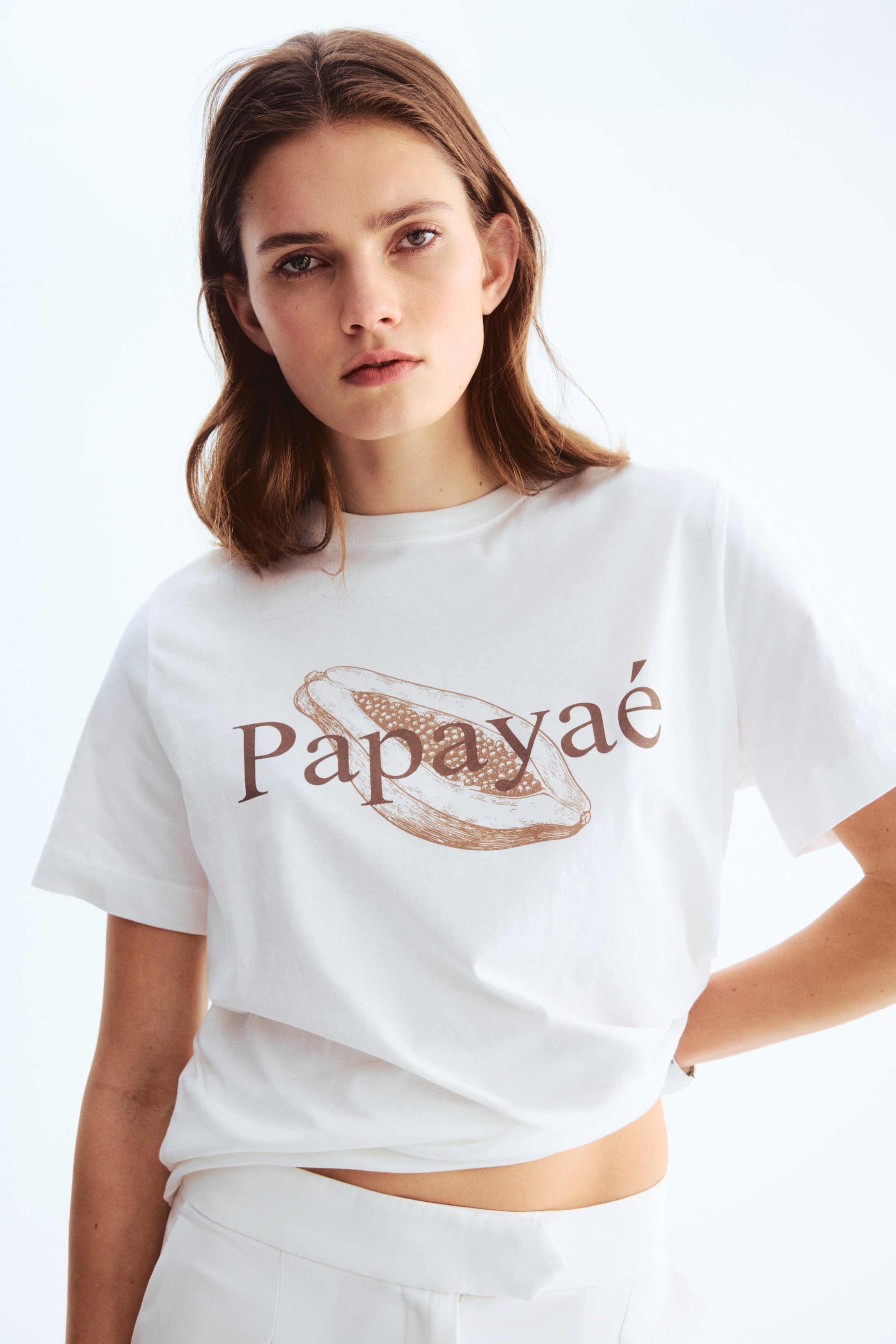 View larger image: A white short-sleeved t-shirt showcases a round neckline and a sepia-toned graphic of a halved papaya fruit with the word "Papaya" printed across the front. It is styled with a relaxed fit, partially tucked into white trousers at the front.