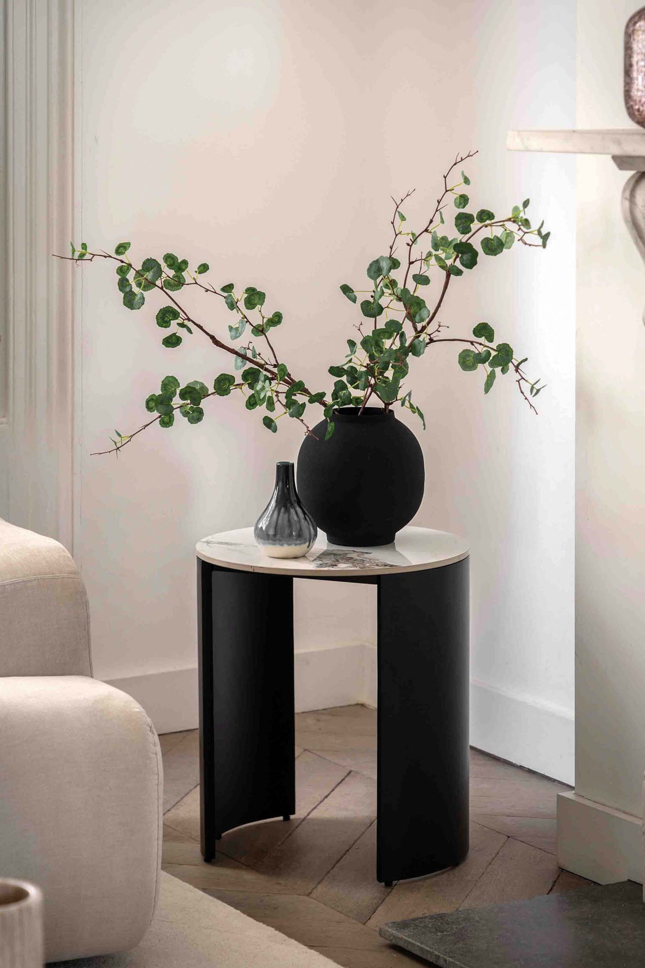 View larger image: A side table featuring a round, light-toned marble top with subtle grey veining, supported by a black base formed by two curved, black panel legs. A large matte black spherical vase holding green branches and a smaller grey and white teardrop-shaped vase rest on its surface.