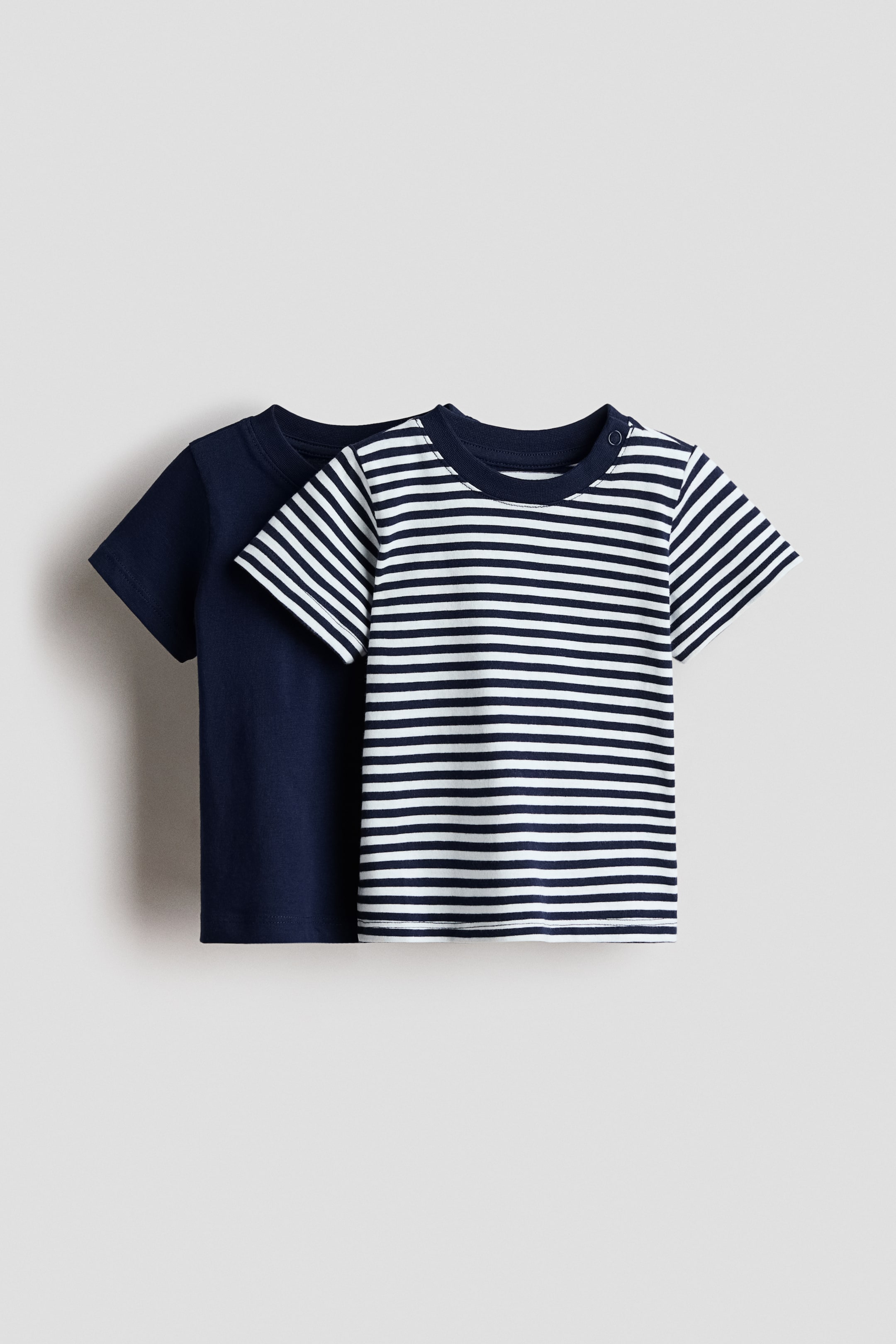 Visualizza immagine più grande: Un pacco da 2 di T-shirt in cotone mostra i suoi lati frontali, con una T-shirt blu navy a tinta unita posizionata dietro una T-shirt bianca con righe orizzontali blu navy. Entrambe le T-shirt presentano maniche corte e scollature rotonde, e la T-shirt a righe include anche bottoni automatici sulla spalla sinistra.