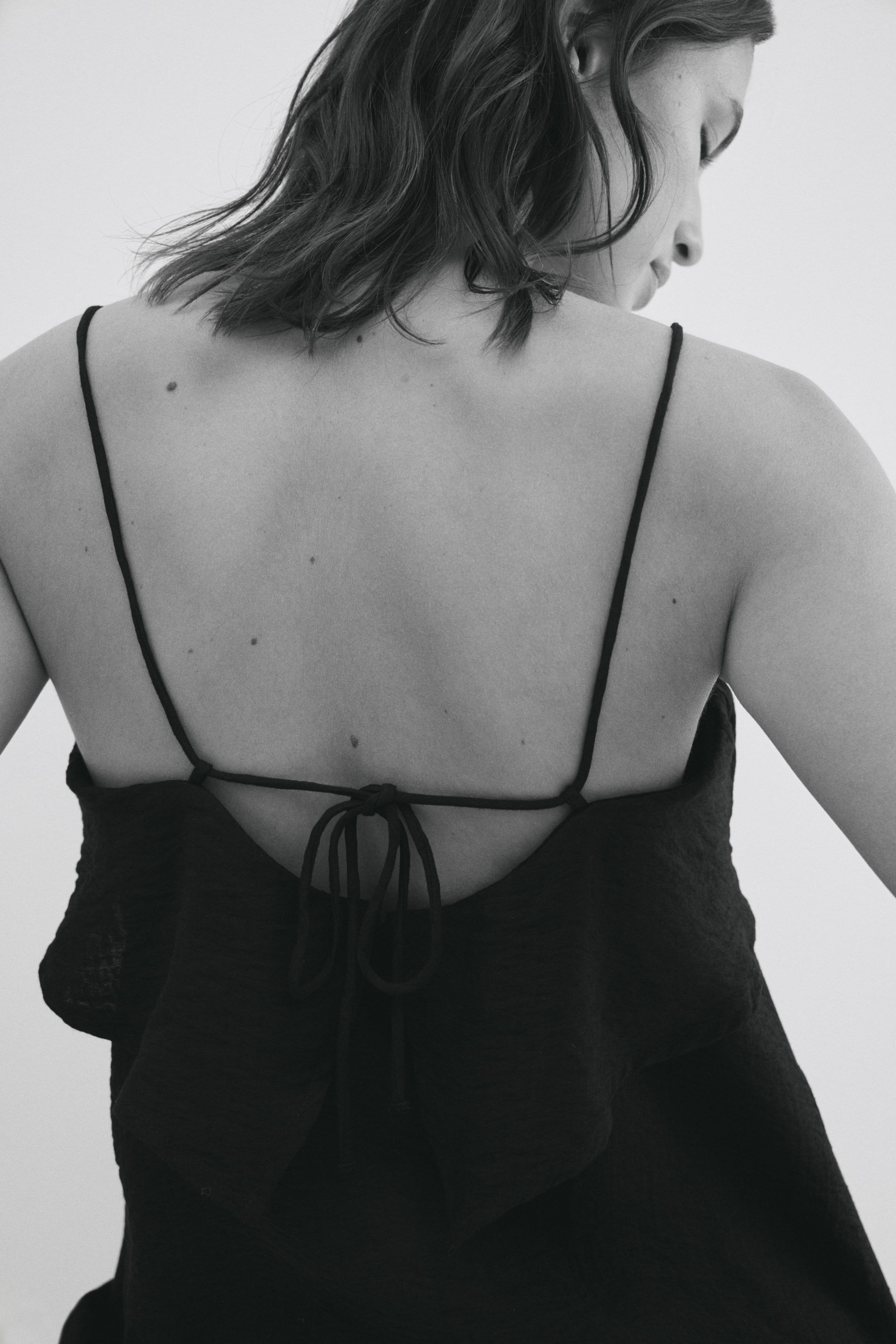View larger image: Black top, viewed from the back, showcases thin spaghetti straps that crisscross and tie into a bow, forming an open back design. The fabric of the garment exhibits a subtle crinkled texture.