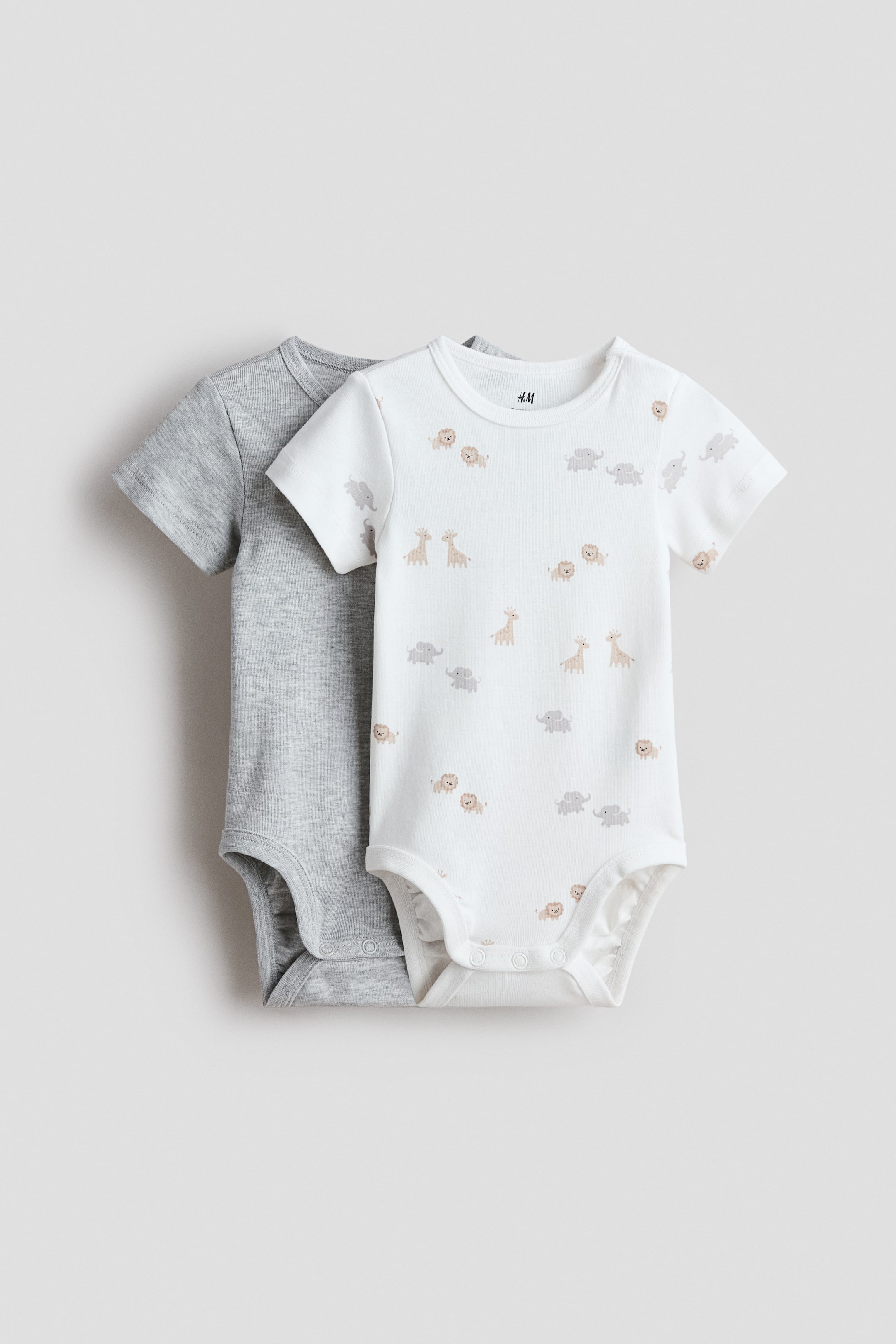 View larger image: Two short-sleeve bodysuits are displayed, both facing forward. The front bodysuit is white, showcasing a repeating pattern of small giraffes, elephants, and lions, with a trimmed round neckline and snap fastenings at the bottom. The bodysuit behind it is a solid light gray and also features a trimmed round neckline.
