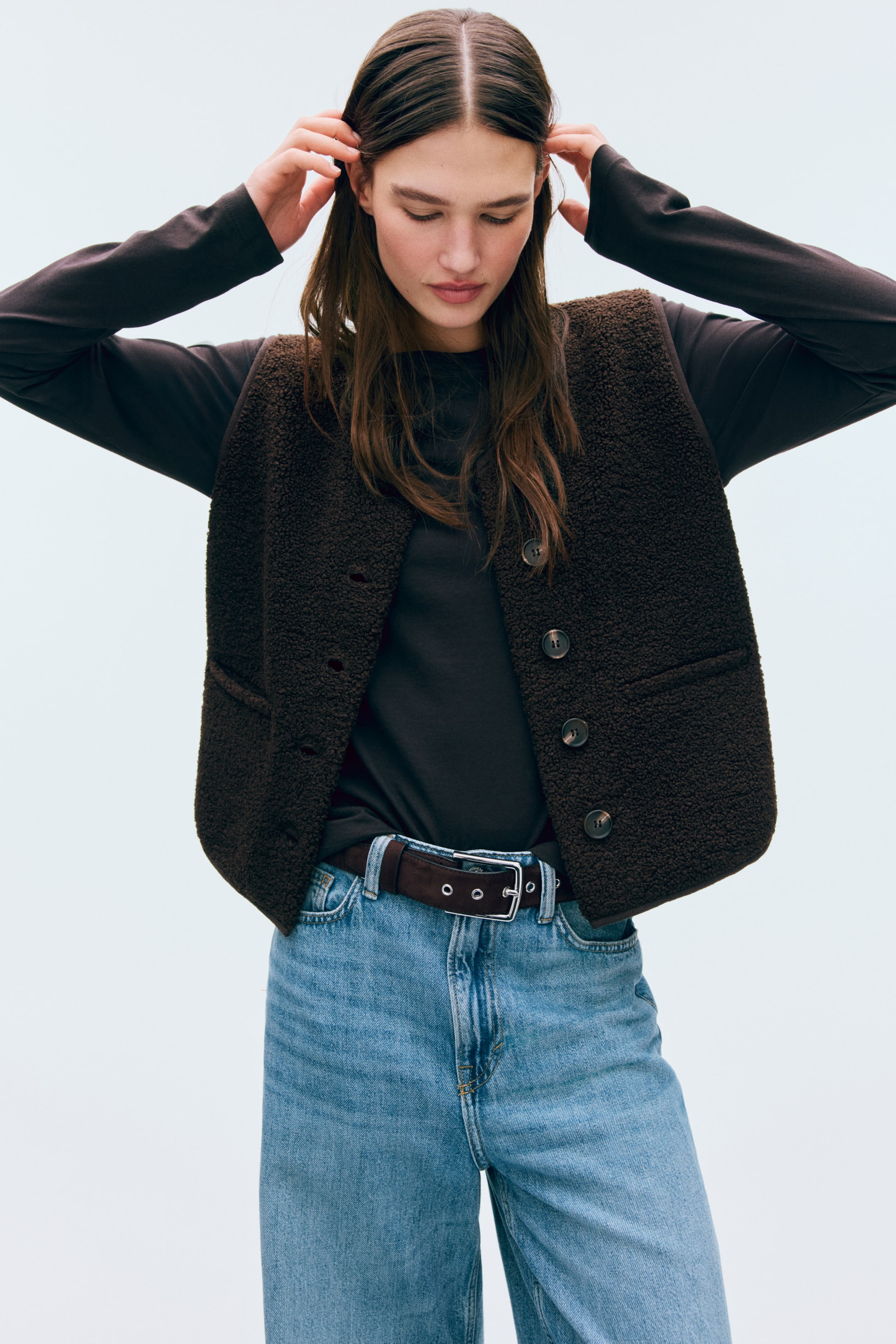 Größeres Bild anzeigen: Eine dunkelbraune, strukturierte Weste wird offen über einem dunklen Langarmoberteil getragen und ist mit vier vorn sichtbaren Knöpfen sowie zwei aufgesetzten Taschen ausgestattet.