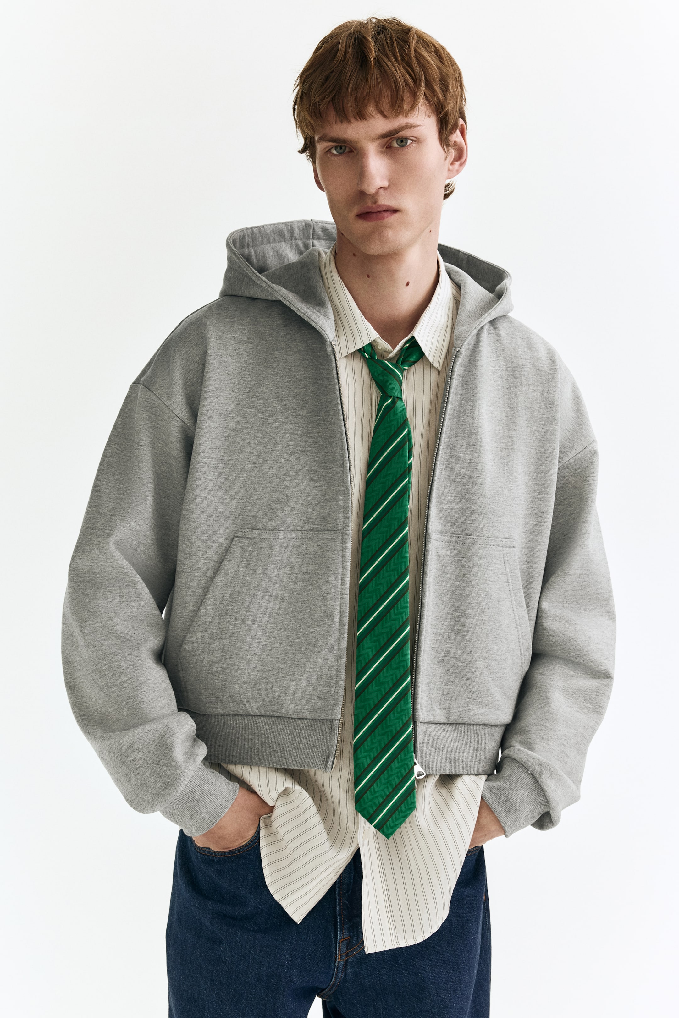 拡大画像を表示: グレーのジップアップフーディーが着用されています。フルジップのフロント、付属のフード、そして二つのフロントポケットが特徴です。リラックスしたクロップドフィットで、リブ付きの袖口と裾が施されています。ストライプのシャツとグリーンのネクタイの上にレイヤードされています。