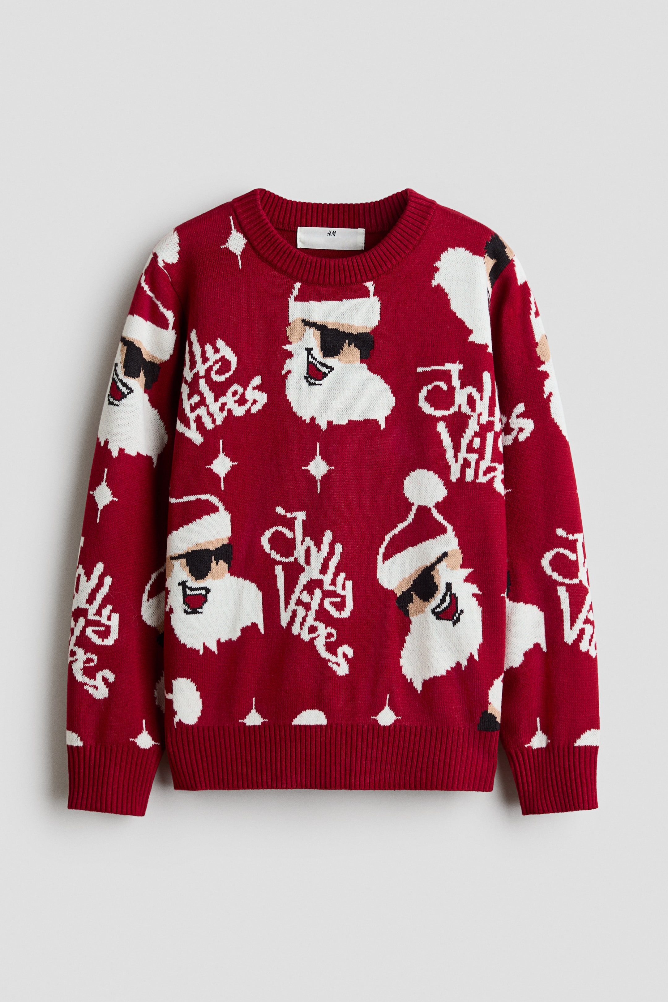 View larger image: A red long-sleeved crew-neck sweater is shown from the front, featuring a repeating knit pattern of Santa Claus wearing sunglasses, stylised white script text reading "Jolly Vibes", and small white star motifs. The garment showcases a ribbed neckline, ribbed cuffs, and a ribbed hem.