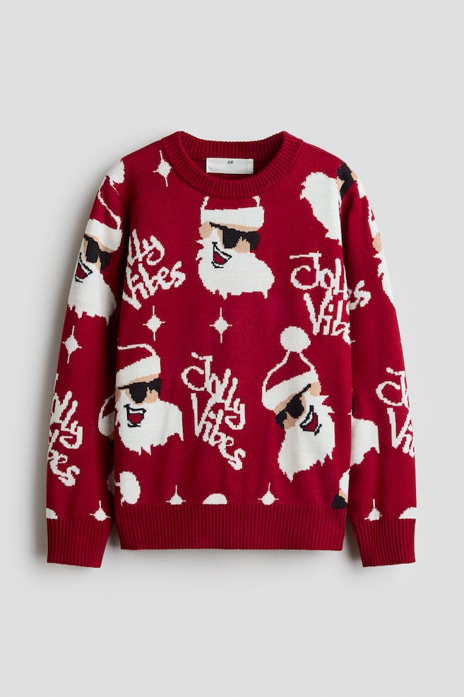 A red long-sleeved crew-neck sweater is shown from the front, featuring a repeating knit pattern of Santa Claus wearing sunglasses, stylised white script text reading "Jolly Vibes", and small white star motifs. The garment showcases a ribbed neckline, ribbed cuffs, and a ribbed hem. - 1