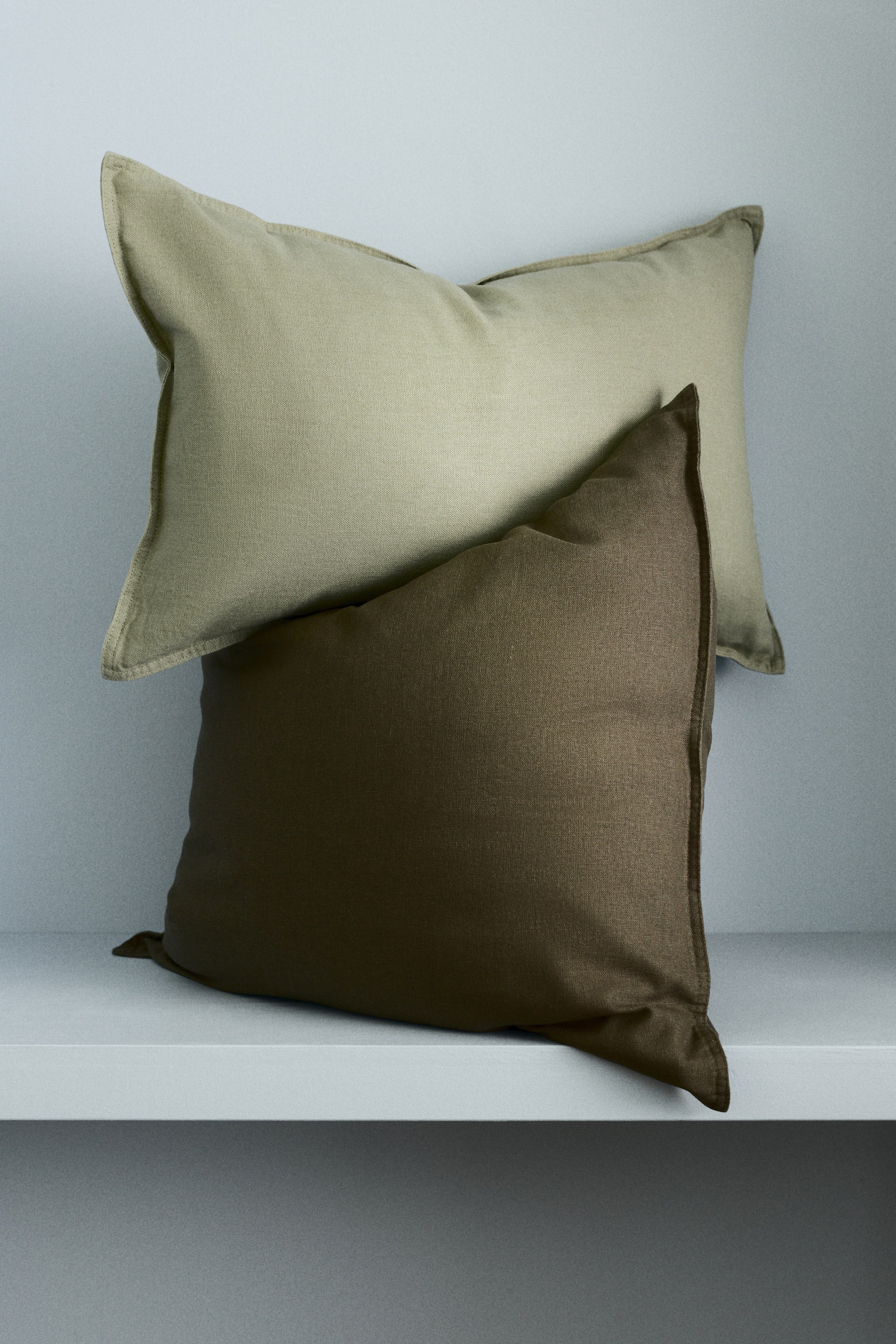 View larger image: A cushion cover in a light beige linen and cotton weave is resting on a similar cushion cover in dark olive green. Both cushion covers are displaying stitching along their sides.