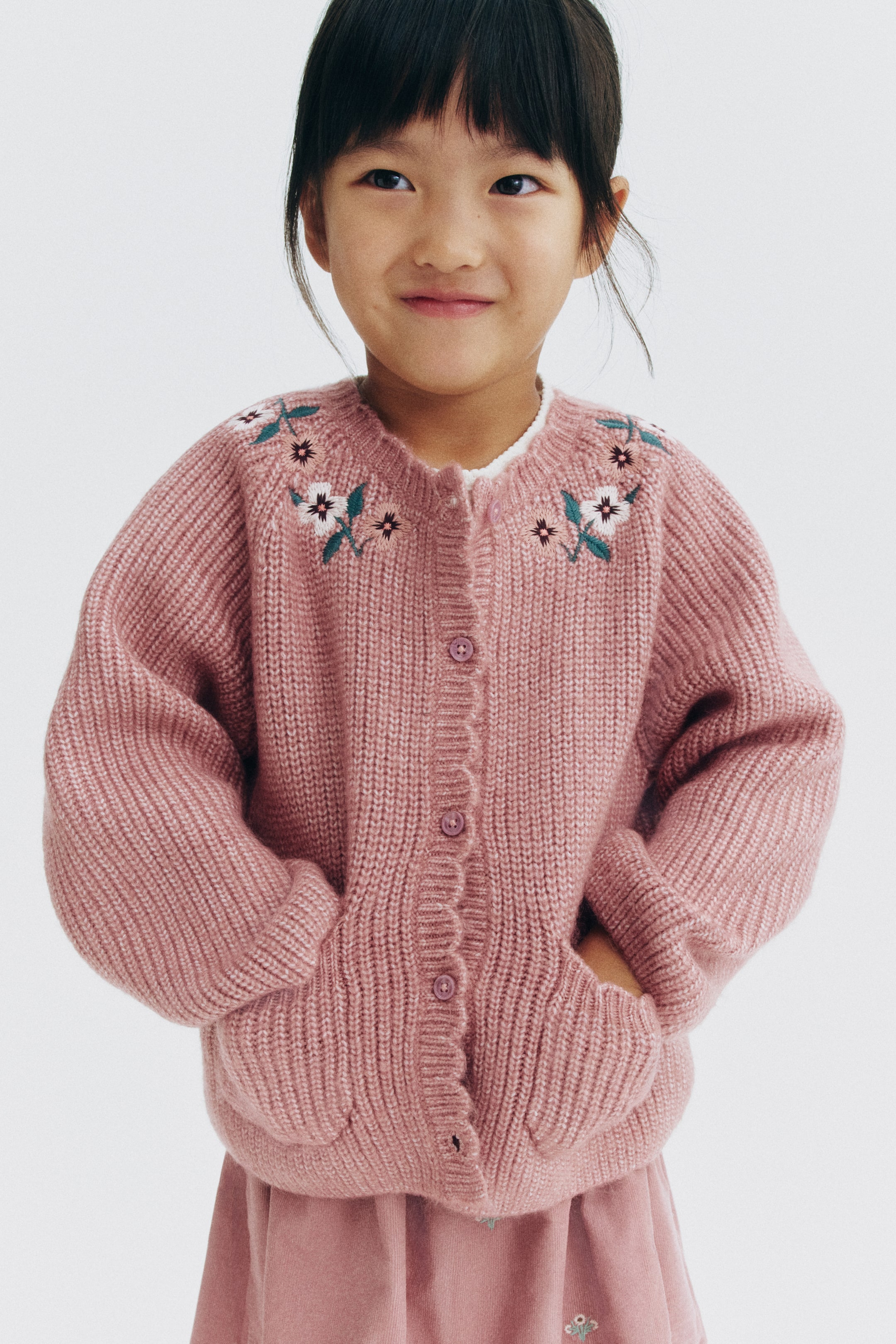View larger image: A dusty pink cardigan in a soft English rib knit is shown facing front, featuring delicate floral embroidery on the shoulders and a round, scalloped neckline. The cardigan is displaying a button-down front, open patch pockets, and ribbed cuffs, layered over a white shirt.