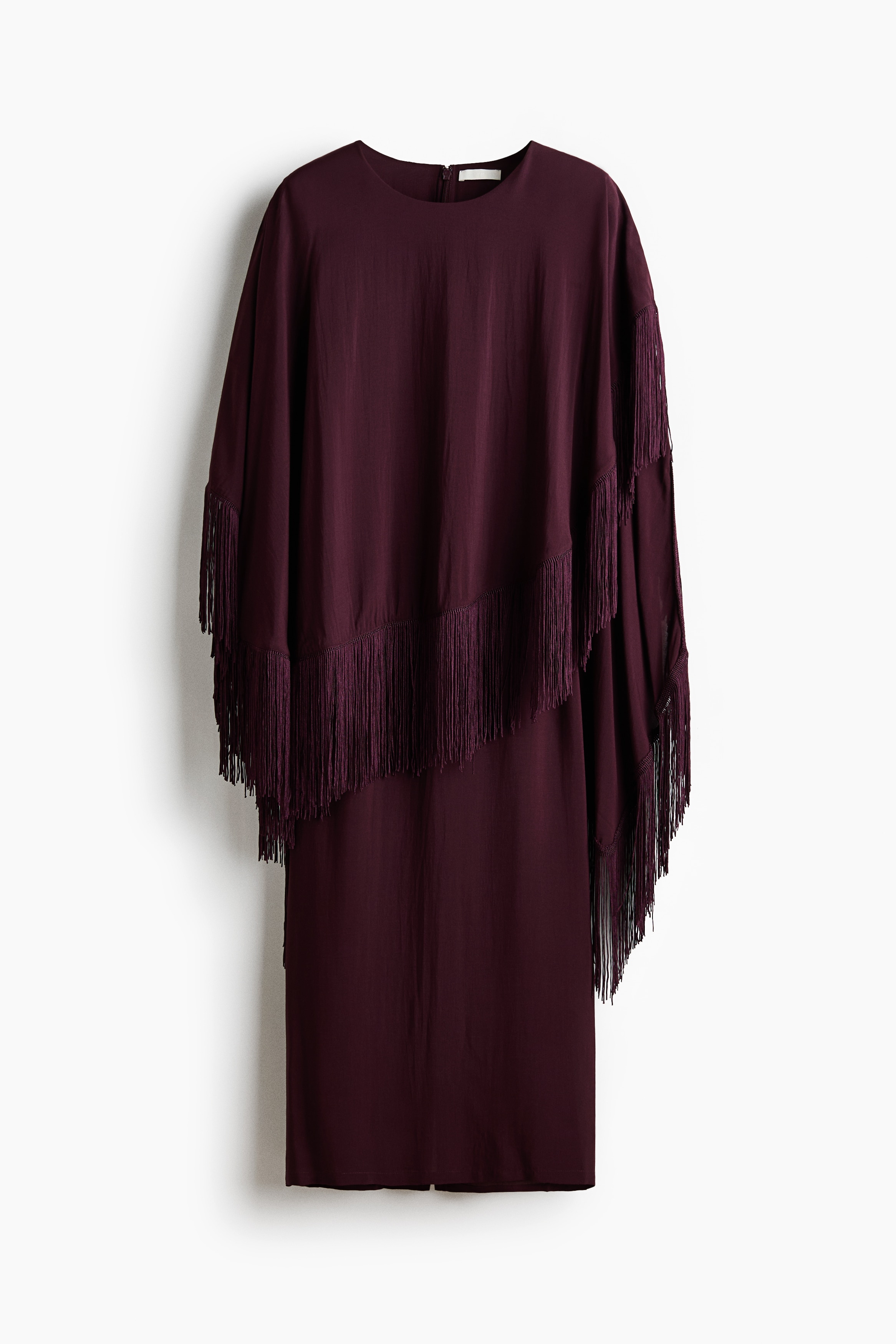 The model is wearing a dark plum-coloured long dress, featuring a draped, asymmetrical cape layer with long fringe trim along the bottom edge. The flowing cape layer drapes over the shoulders and upper body, while the straight-cut skirt of the dress is visible below.