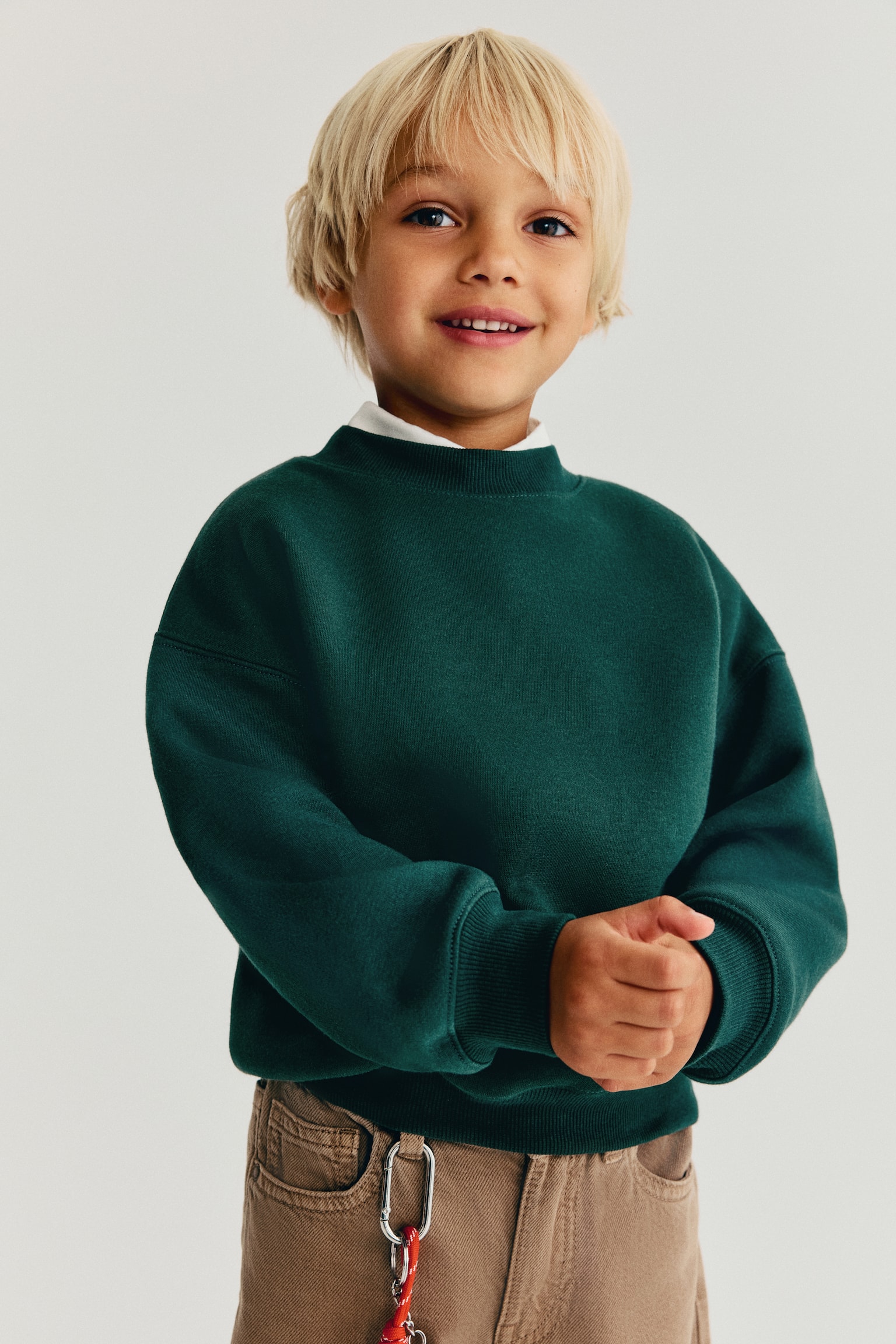 A dark green sweatshirt is shown from the front, featuring a round rib-trimmed neckline, dropped shoulders, and long sleeves with wide ribbed cuffs. The sweatshirt is layered over a white collar and finished with a ribbed hem. - 1