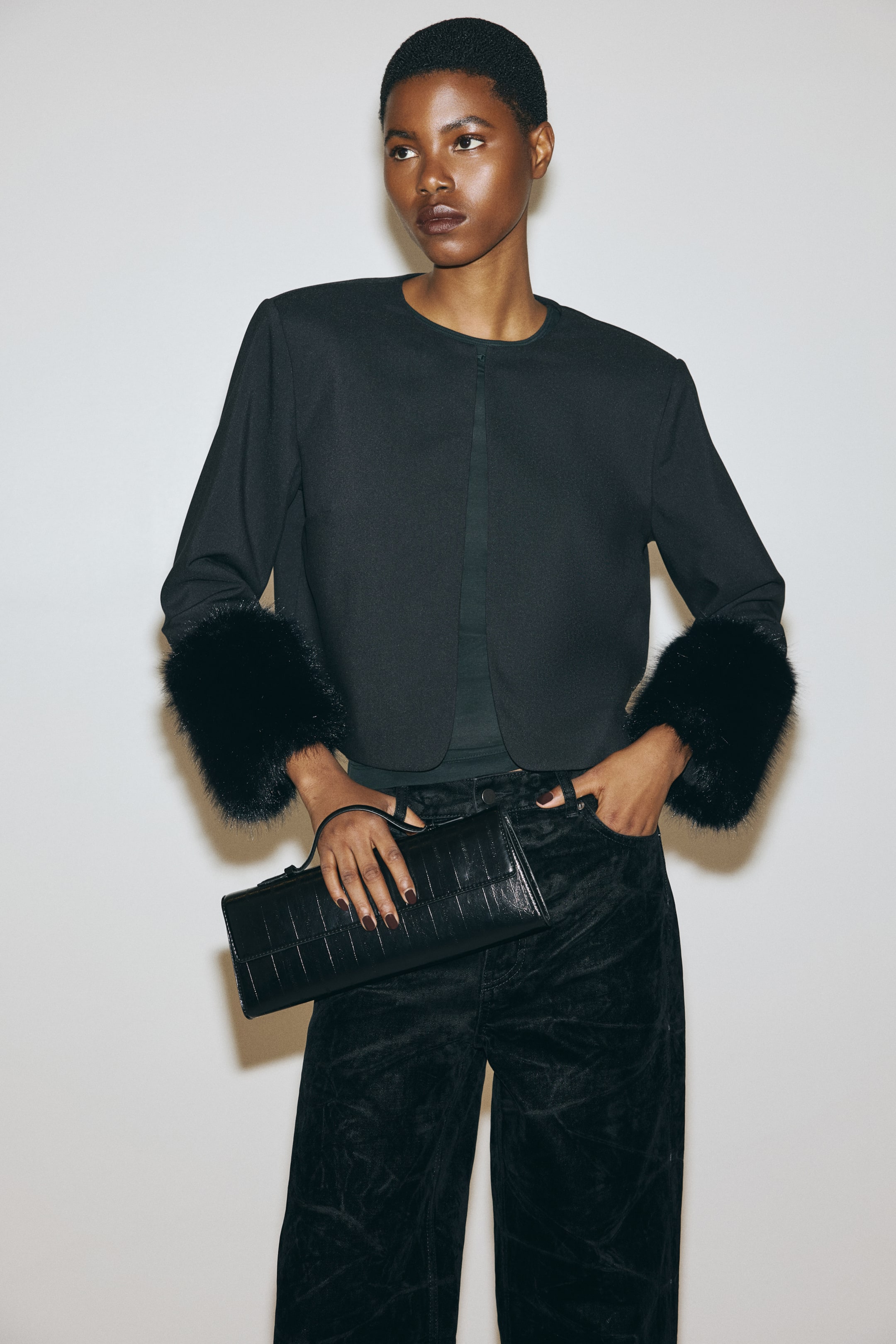 View larger image: A black cropped jacket is being presented from the front. It features a round, collarless neckline, an open front, and long sleeves ending in prominent black fur cuffs.