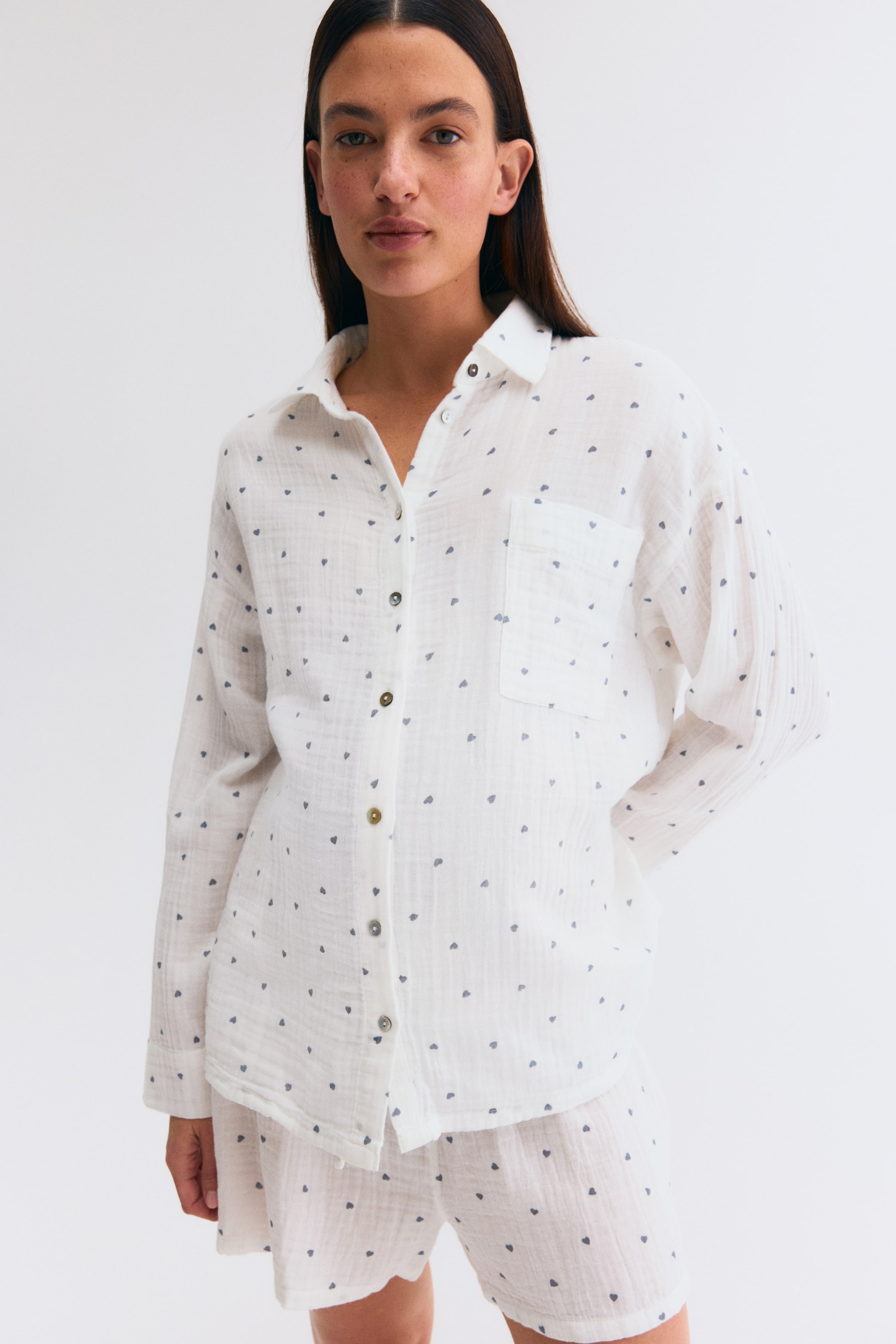 Ver imagen más grande: Se muestra un conjunto de pijama blanco visto desde el frente, con un estampado de pequeños corazones grises por toda la prenda. La camisa de manga larga con cuello presenta un bolsillo en el pecho y botones marrones, con algunos botones actualmente desabrochados. Los pantalones cortos a juego están diseñados con una cinturilla fruncida.
