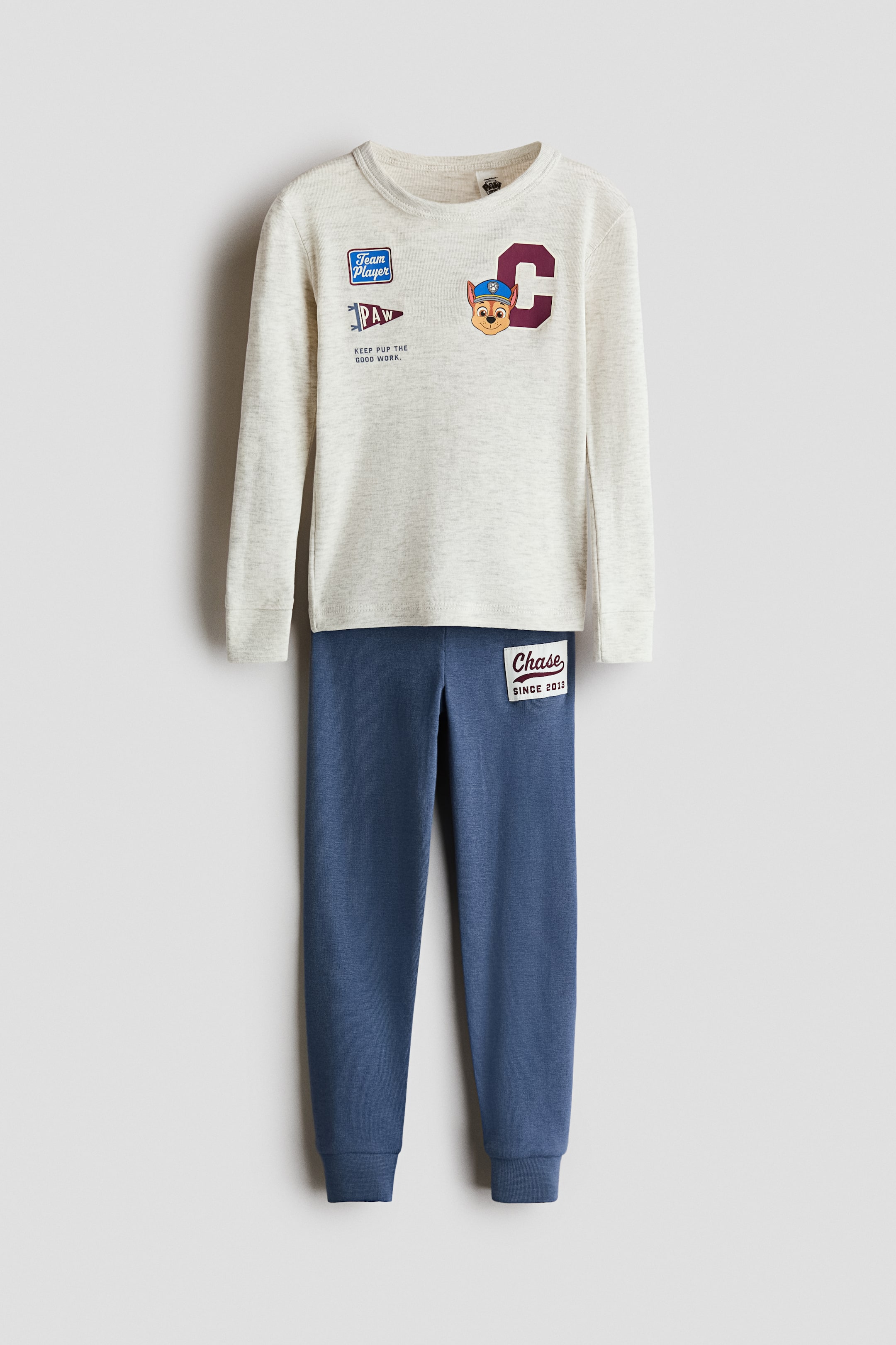 View larger image: A two-piece children's loungewear set is displayed flat, facing forward. The light gray marled long-sleeve top features a 'Team Player' patch, a 'PAW' pennant, and a character head next to a burgundy letter 'C' on the front. The coordinating blue marled pants feature a 'Chase Since 2013' patch on the upper left thigh and ribbed ankle cuffs.