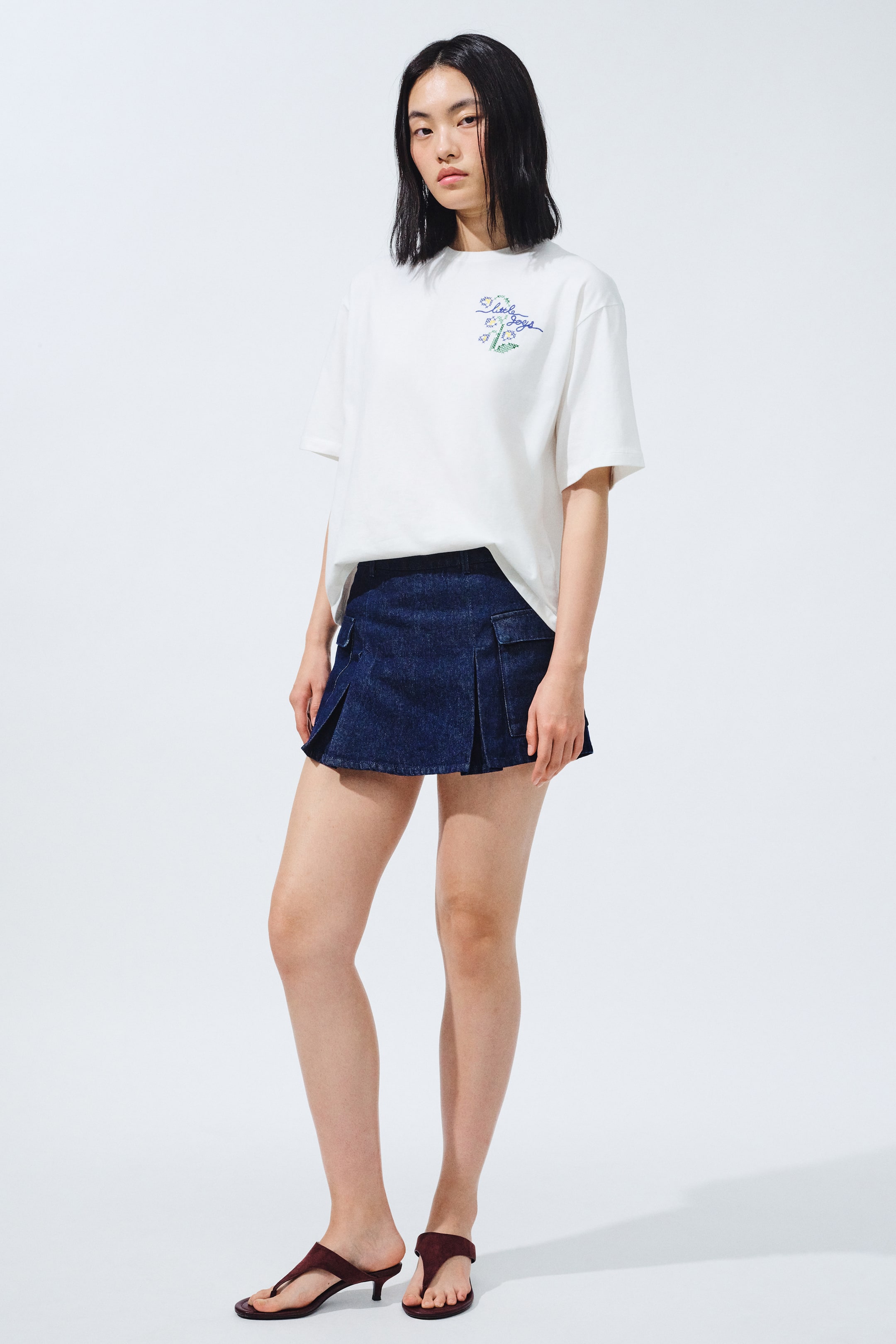 拡大画像を表示: 白の半袖Tシャツは、左胸に刺繍された花柄とテキストデザインが施されており、濃紺のデニムミニスカートは、前面プリーツと両サイドにフラップポケットが付いています。すべてがやや前右からの角度で示されています。