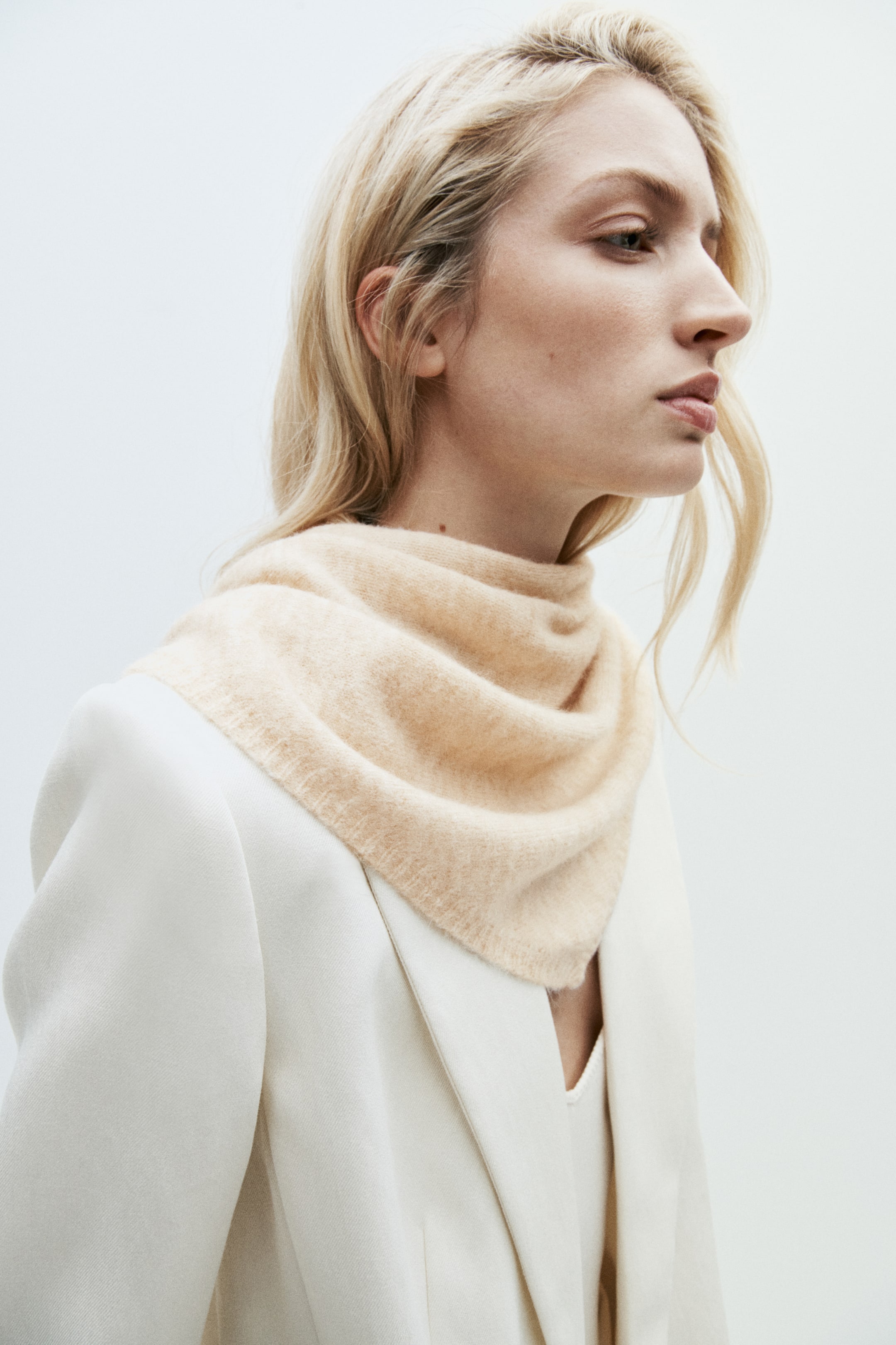 View larger image: A light beige, soft-knit triangle scarf with a narrow ribbed trim is worn draped around the model’s neck. The scarf is layered over a structured white blazer.