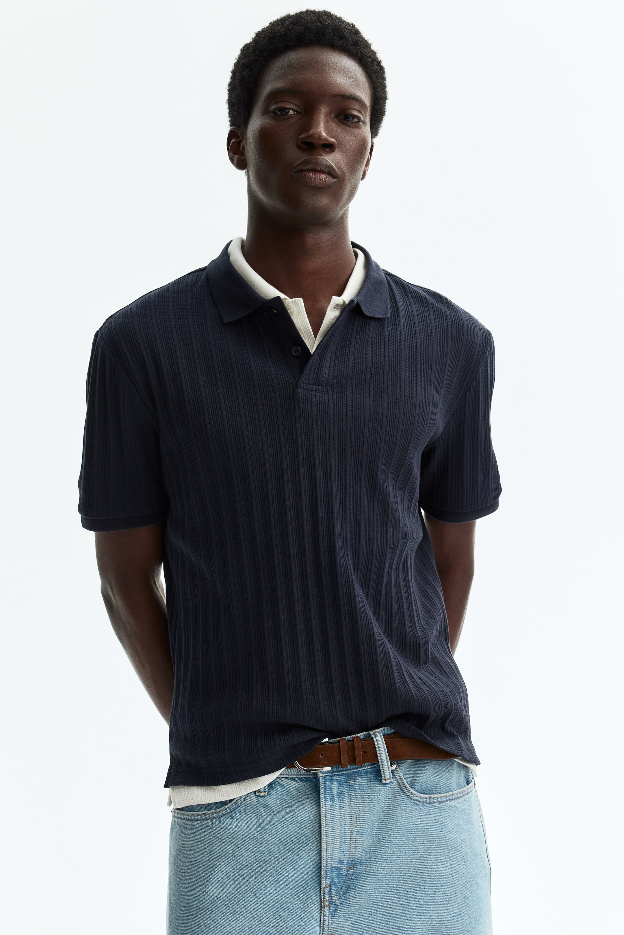 View larger image: The navy blue ribbed polo shirt is displayed front-facing, showcasing a classic collar and a two-button placket. It features a vertical ribbed texture, short sleeves, and a relaxed fit, with a light-coloured undershirt visible beneath.