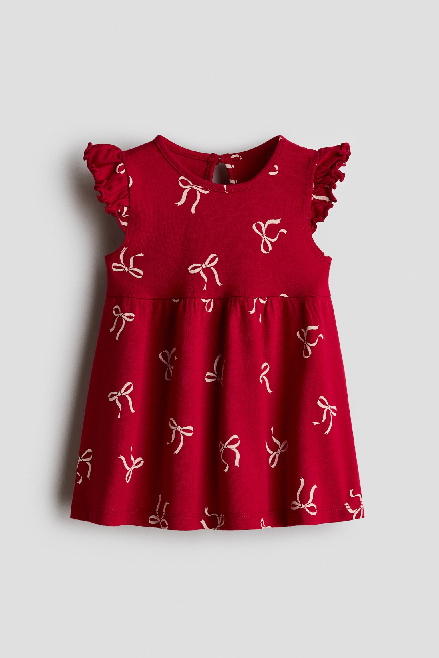 A red cotton jersey dress is shown from the front, featuring an all-over print of small white bows. The dress is designed with a round neckline, ruffled flutter sleeves, gathers at the waist, and a flared skirt.