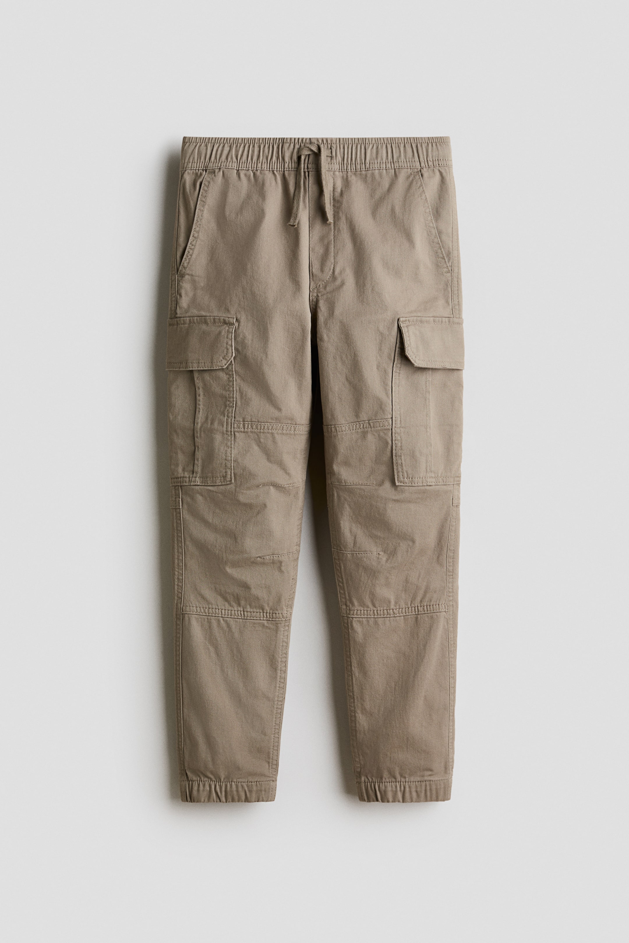 View larger image: A pair of khaki cotton twill cargo joggers is presenting from the front, showcasing a relaxed fit. They are featuring an elasticated drawstring waist with a fake fly, diagonal front pockets, prominent flap cargo pockets on the legs, and covered elastication detailing the hems.