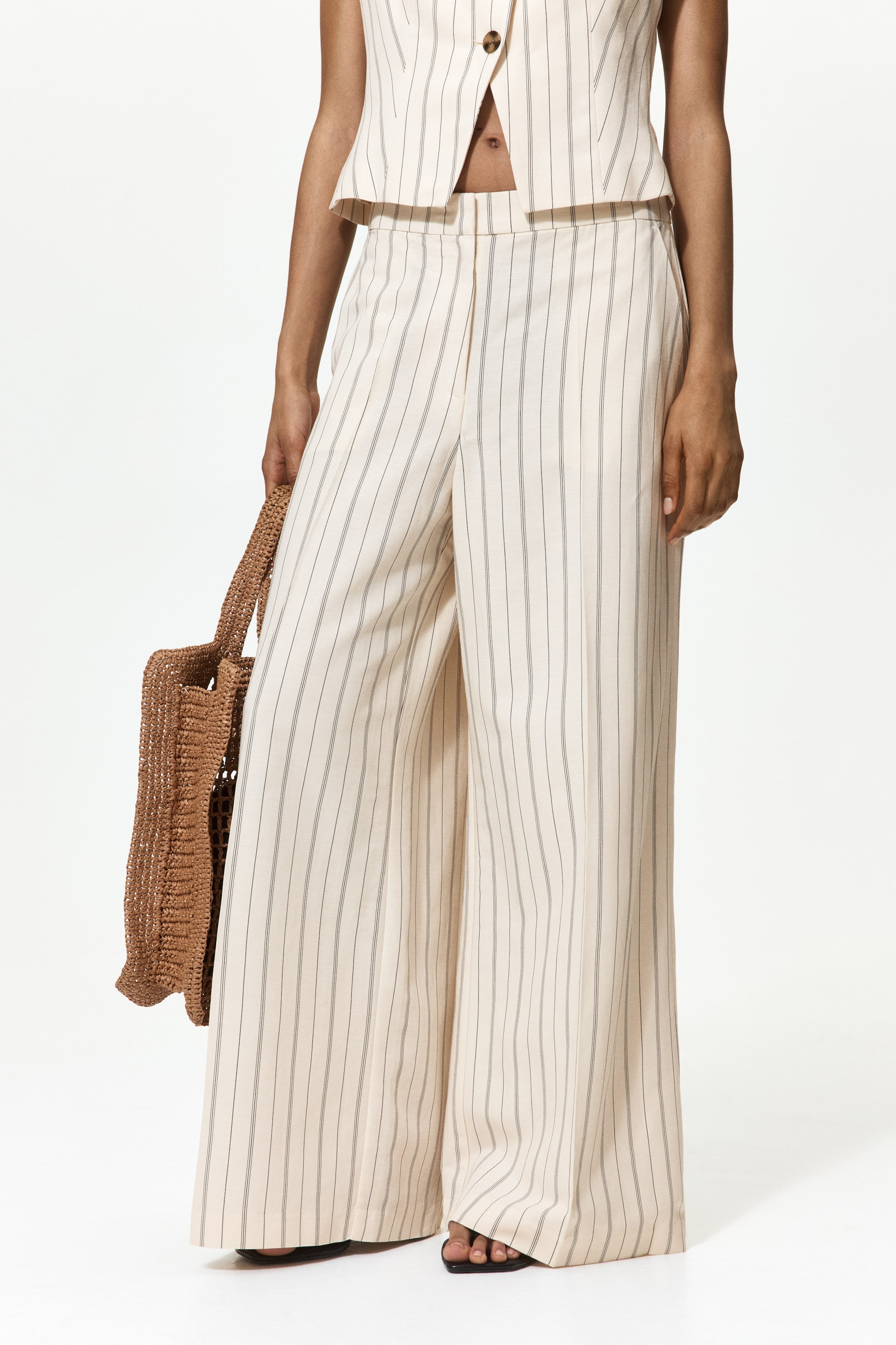 View larger image: Loose-fit, off-white trousers with thin vertical dark pinstripes are designed with a high waist at the front, featuring a zip fly, a concealed hook-and-bar fastener, and a button. They showcase diagonal front pockets and wide legs with prominent creases down the center.