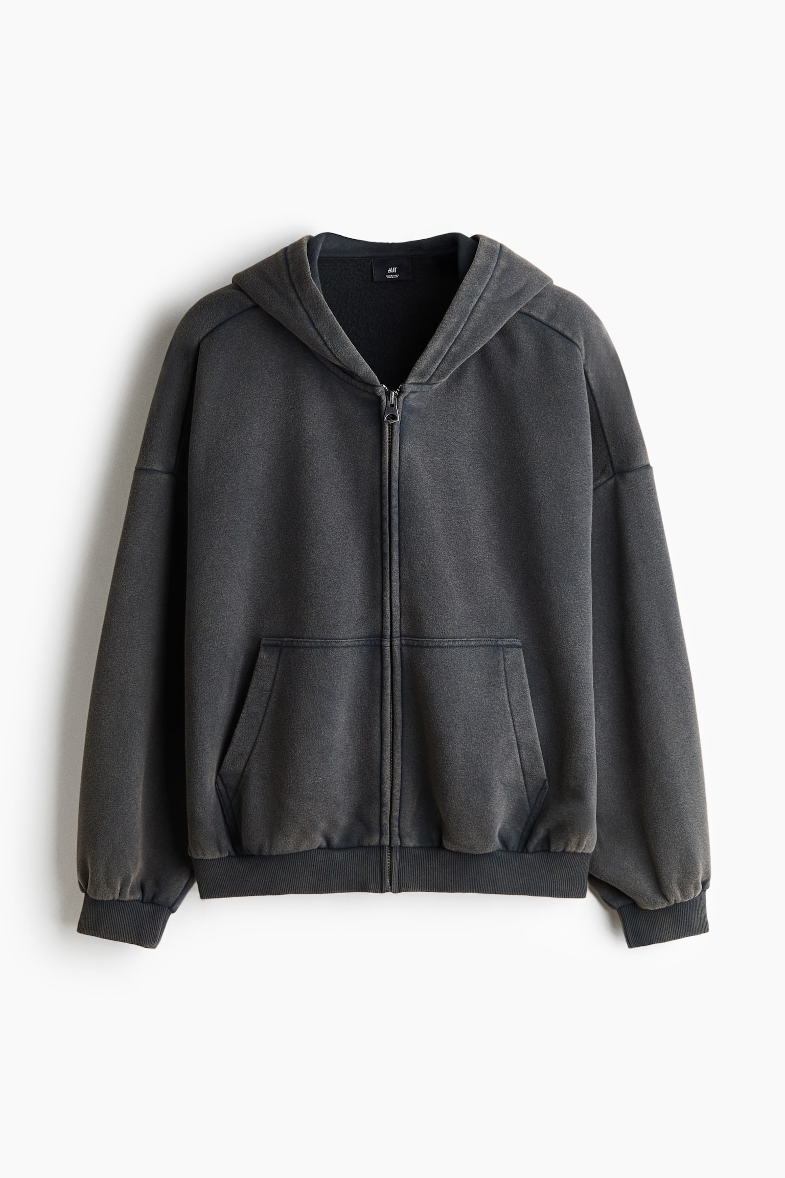 A dark grey zip-up hoodie is shown from the front, featuring a washed-effect finish, dropped shoulders, and a full-length front zip. It includes a split kangaroo pocket, ribbed cuffs, and a ribbed hem.