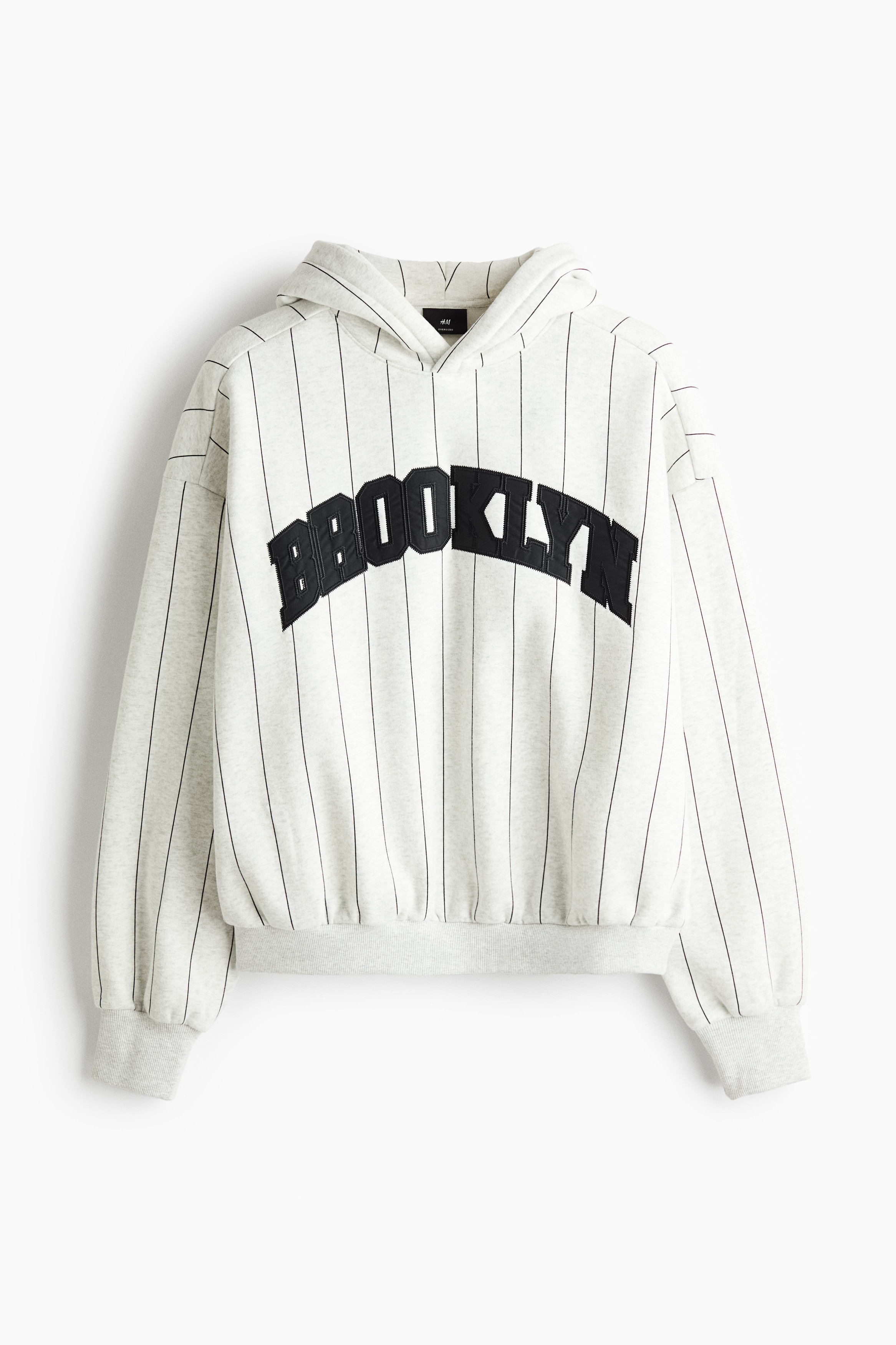A light grey, pinstriped oversized hoodie featuring a black arched "BROOKLYN" applique across the chest is being worn by the model. The hoodie features a wrap hood, dropped shoulders, and ribbed trim, and is styled with matching pinstripe sweatpants.