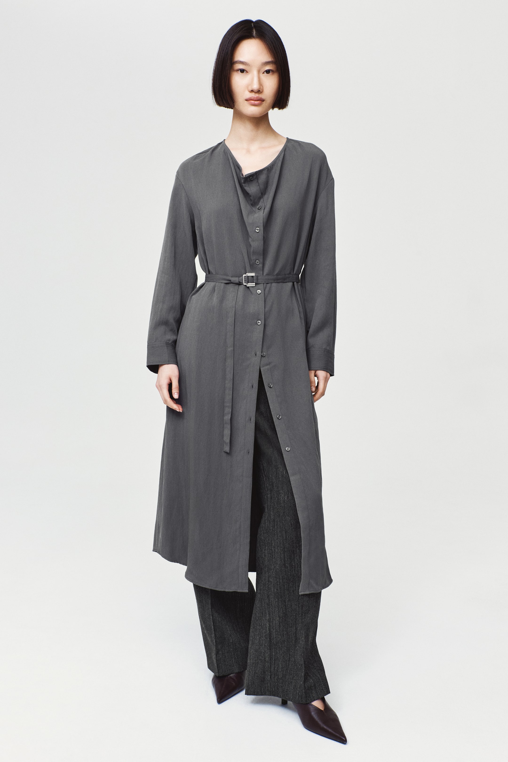 View larger image: A calf-length dark grey dress with a cowl neckline, long sleeves, and an offset button-down front is worn by the model. The dress is cinched at the waist with a slim matching buckle belt and layered over dark wide-leg trousers.