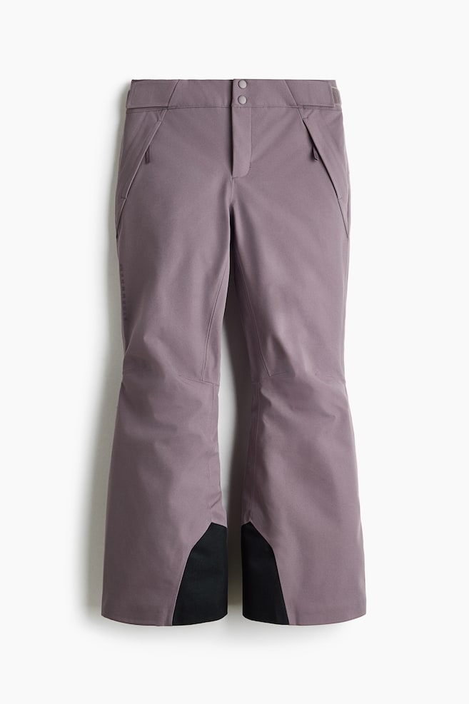 Dusty purple ski trousers are shown from the front, featuring a structured waistband with a double snap-button fastening and angled zipped pockets. The design incorporates articulated knee seams and slightly flared cuffs with contrasting black reinforced inner scuff guards. - 1