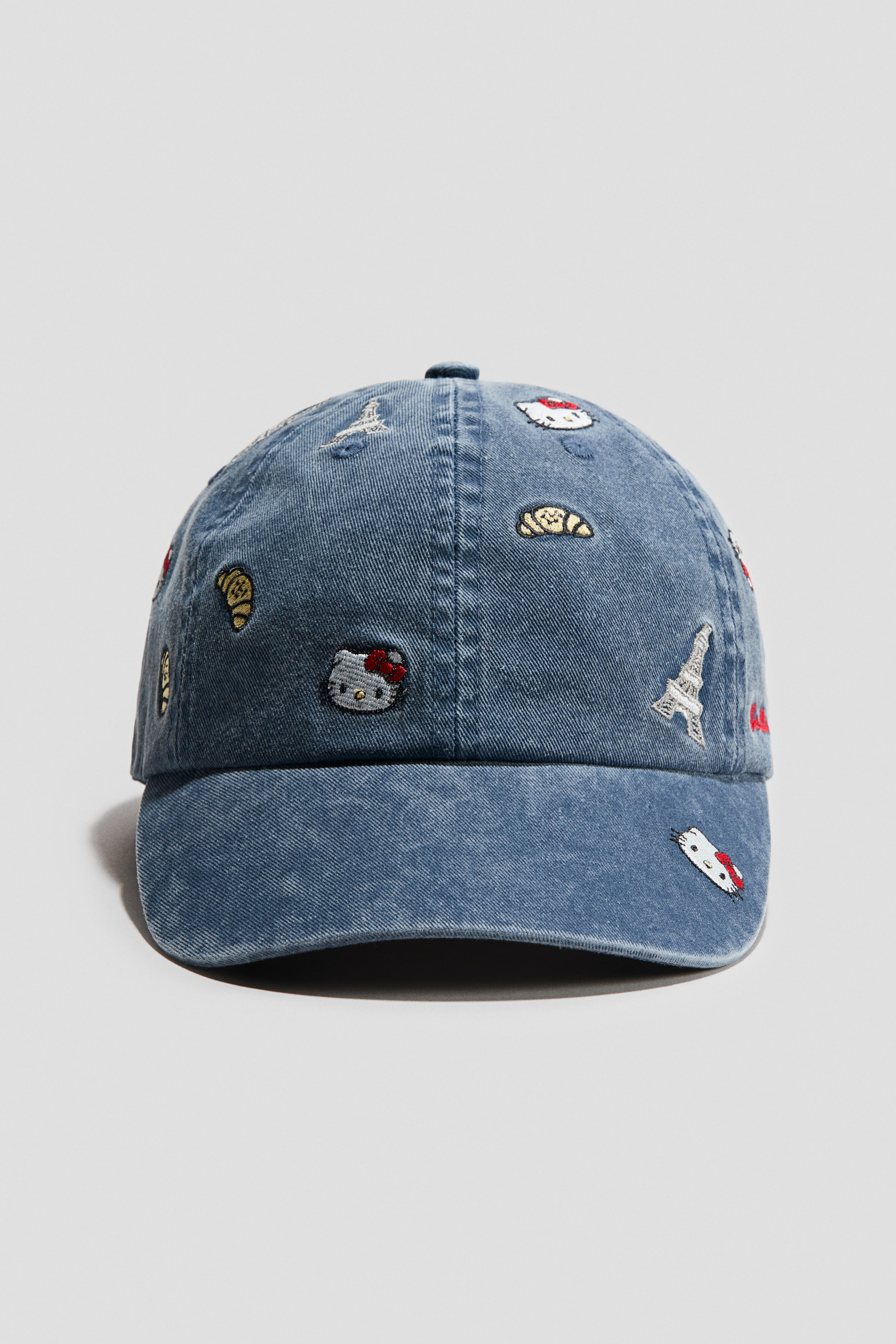 View larger image: The cap is facing FRONT, made of blue faded cotton twill, and is covered with various embroidered motifs including Hello Kitty faces, croissants, the Eiffel Tower, and a bird. Embroidered eyelets are visible on the crown.