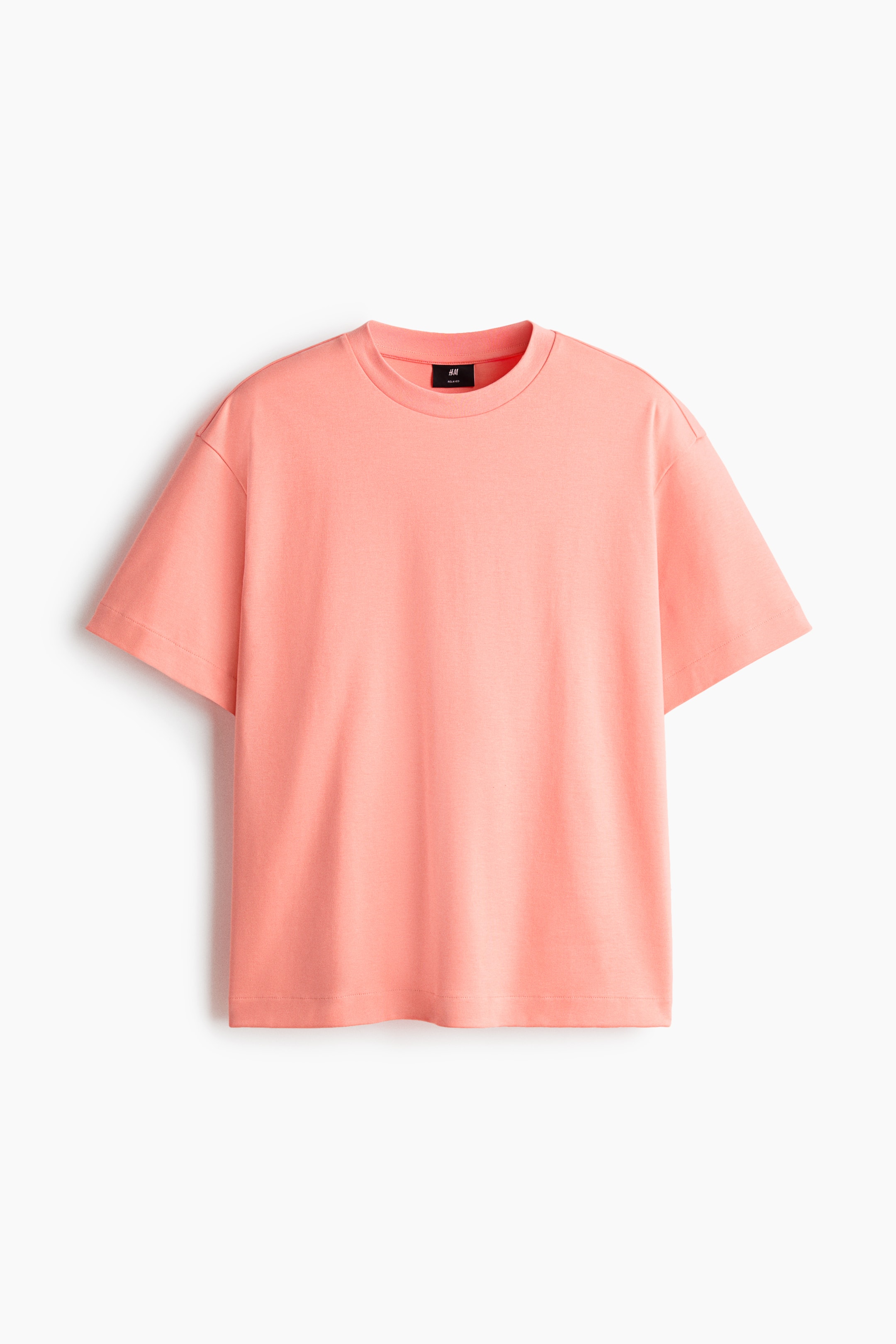 拡大画像を表示: ピーチカラーのボクシースタイルTシャツが正面から見えるように配置されています。丸いリブトリムネックライン、ドロップショルダー、ストレートカットの裾が特徴で、ゆったりとしたフィット感があります。