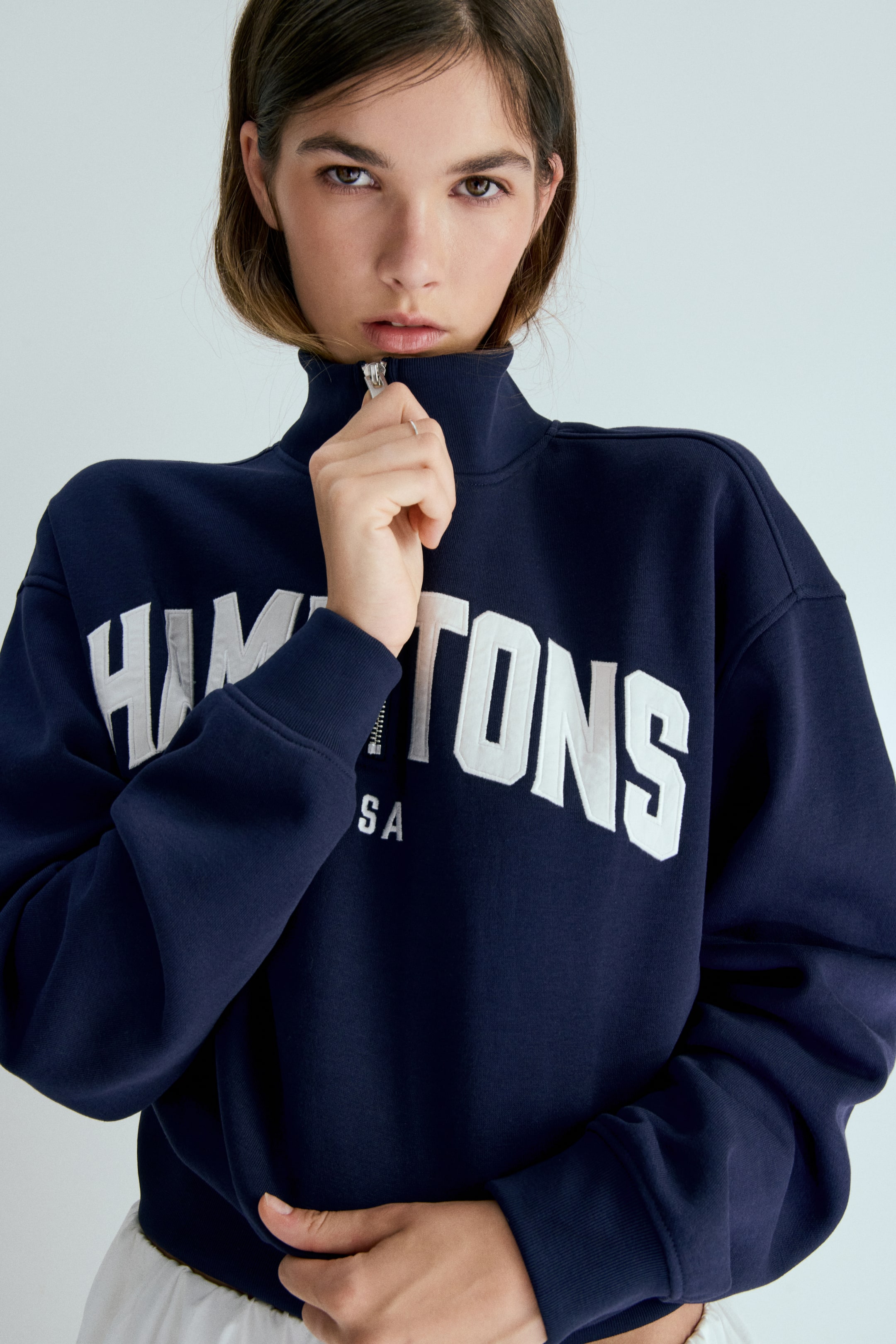 הצגת תמונה גדולה יותר: חולצת סווטשירט בצבע כחול כהה עם רוכסן רבע נלבשת, עם צווארון גבוה ועיטור רוכסן בגוון כסף. רקמה גדולה בלבן מאייתת את המילה "HAMPTONS" על החזה, עם טקסט חלקי "SA" מתחתיה, ושרוולים עם קצוות מצולעים.