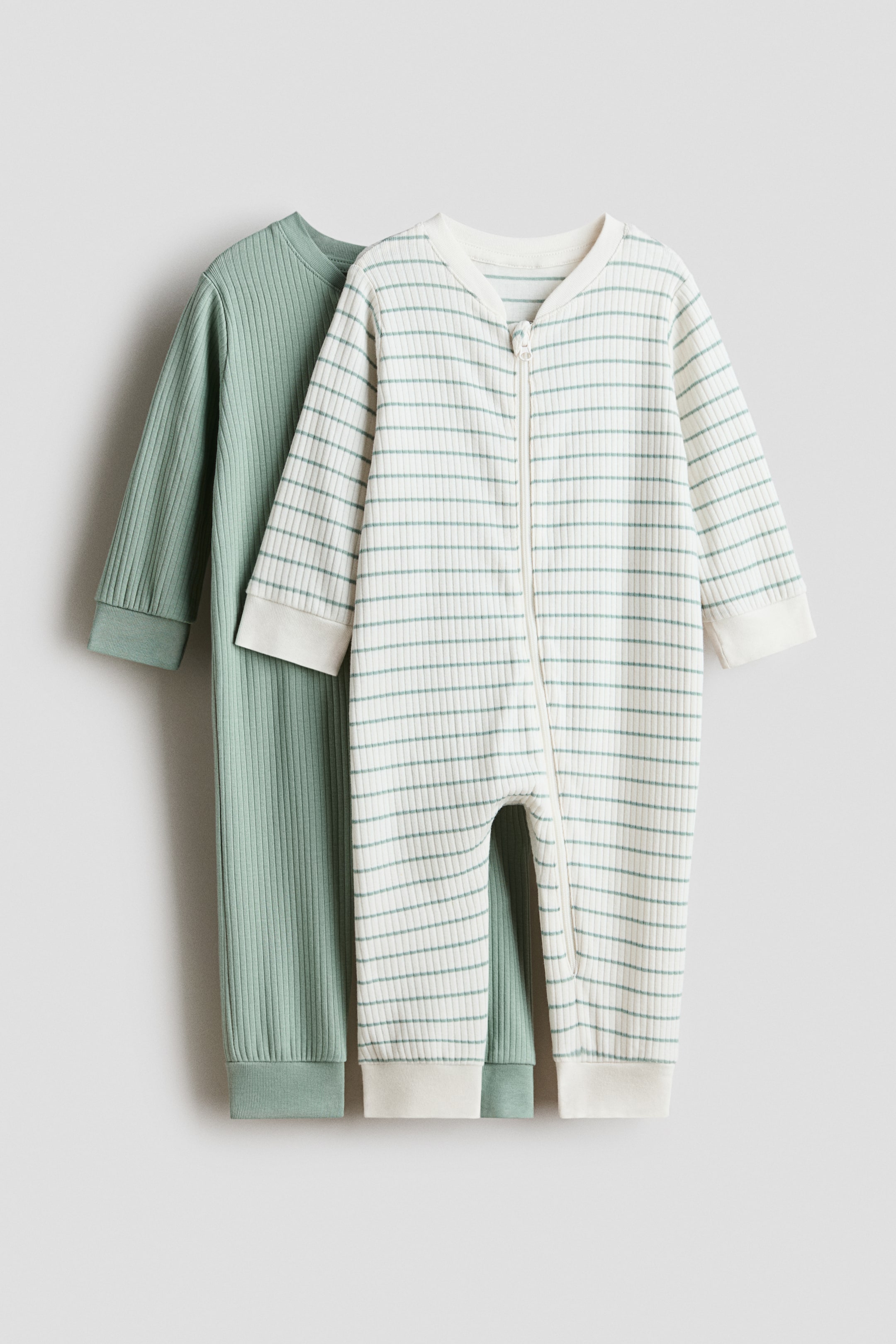 View larger image: Two ribbed sleepsuits are on display: one off-white with horizontal sage-green stripes and one solid sage green. The front striped sleepsuit features a full-length front zipper, long sleeves, and wide ribbed cuffs and ankle trims, with the solid sage-green sleepsuit positioned behind it, also with long sleeves and wide ribbed cuffs and ankle trims.