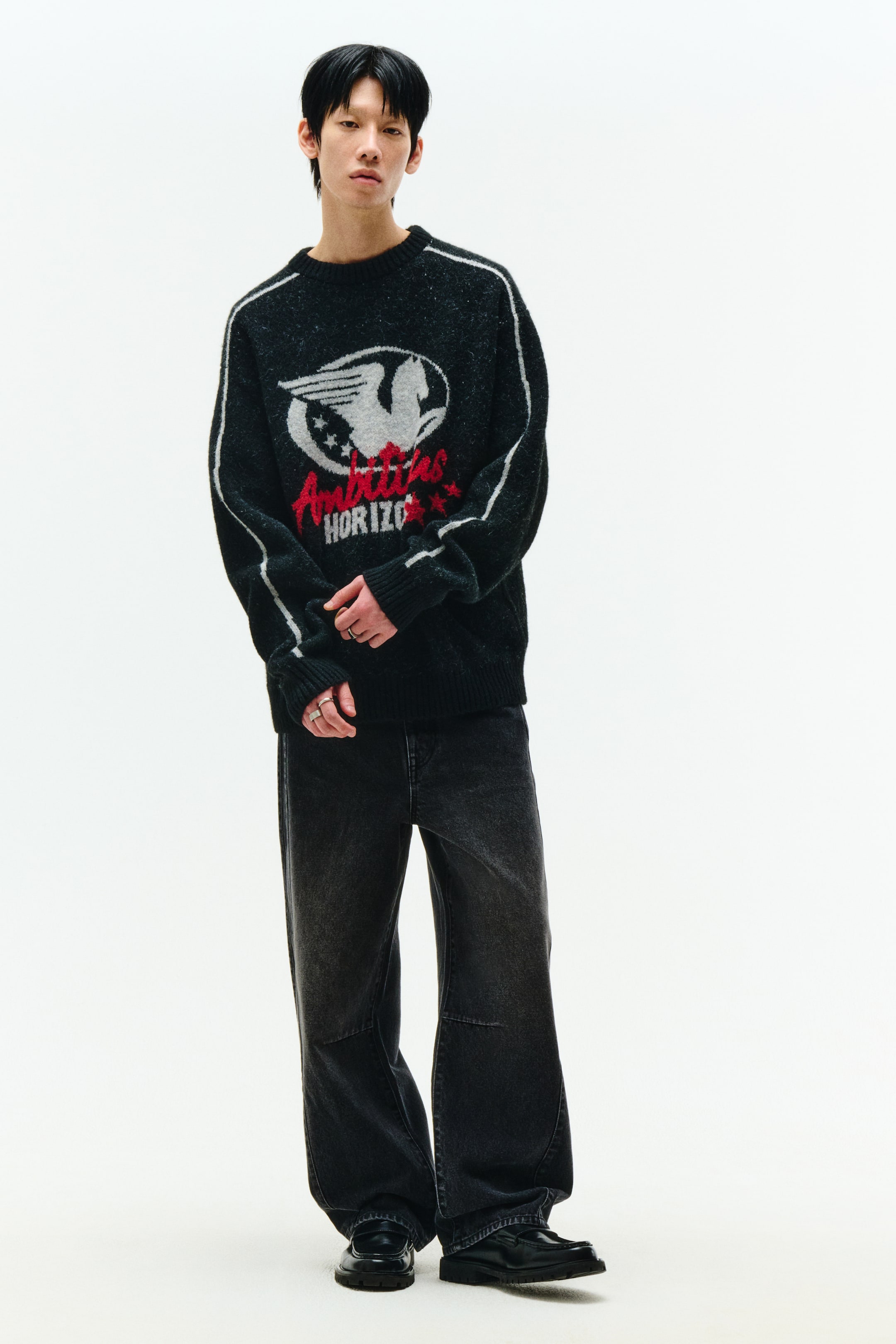 拡大画像を表示: ダークグレーのふわふわしたニットセーターには、フロントに赤い『Ambitious HORIZON』の文字と共に、グレーと白の大きな翼のグラフィックが特色です。両袖には白いストライプが入っており、セーターにはリブ付きのカフスとリブ付きの裾が含まれています。