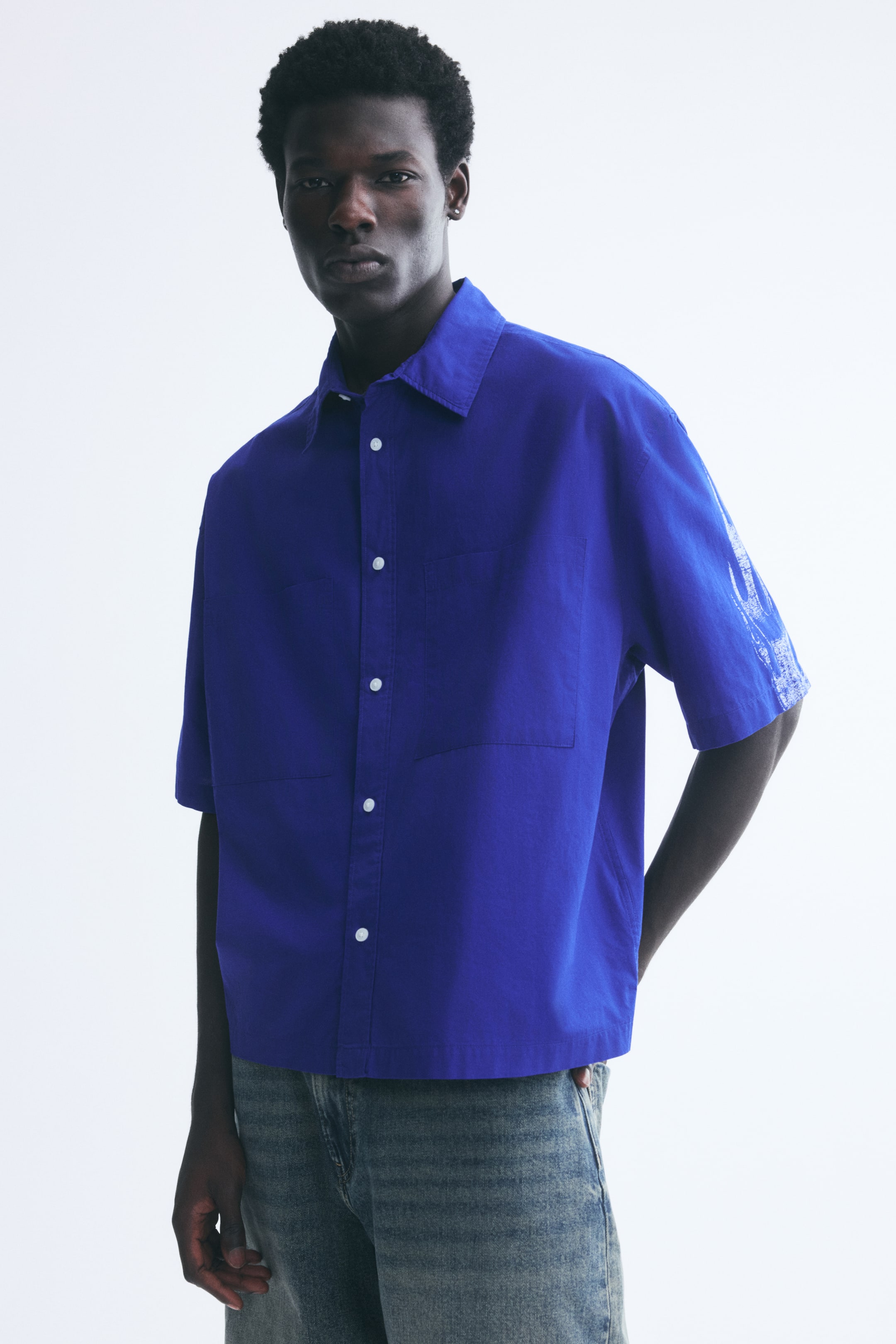 View larger image: A bright blue, short-sleeve button-up shirt is worn by the model, featuring a relaxed fit, a classic collar, dropped shoulders, and patch chest pockets. The shirt features a white printed graphic on the left sleeve and is paired with faded blue jeans.