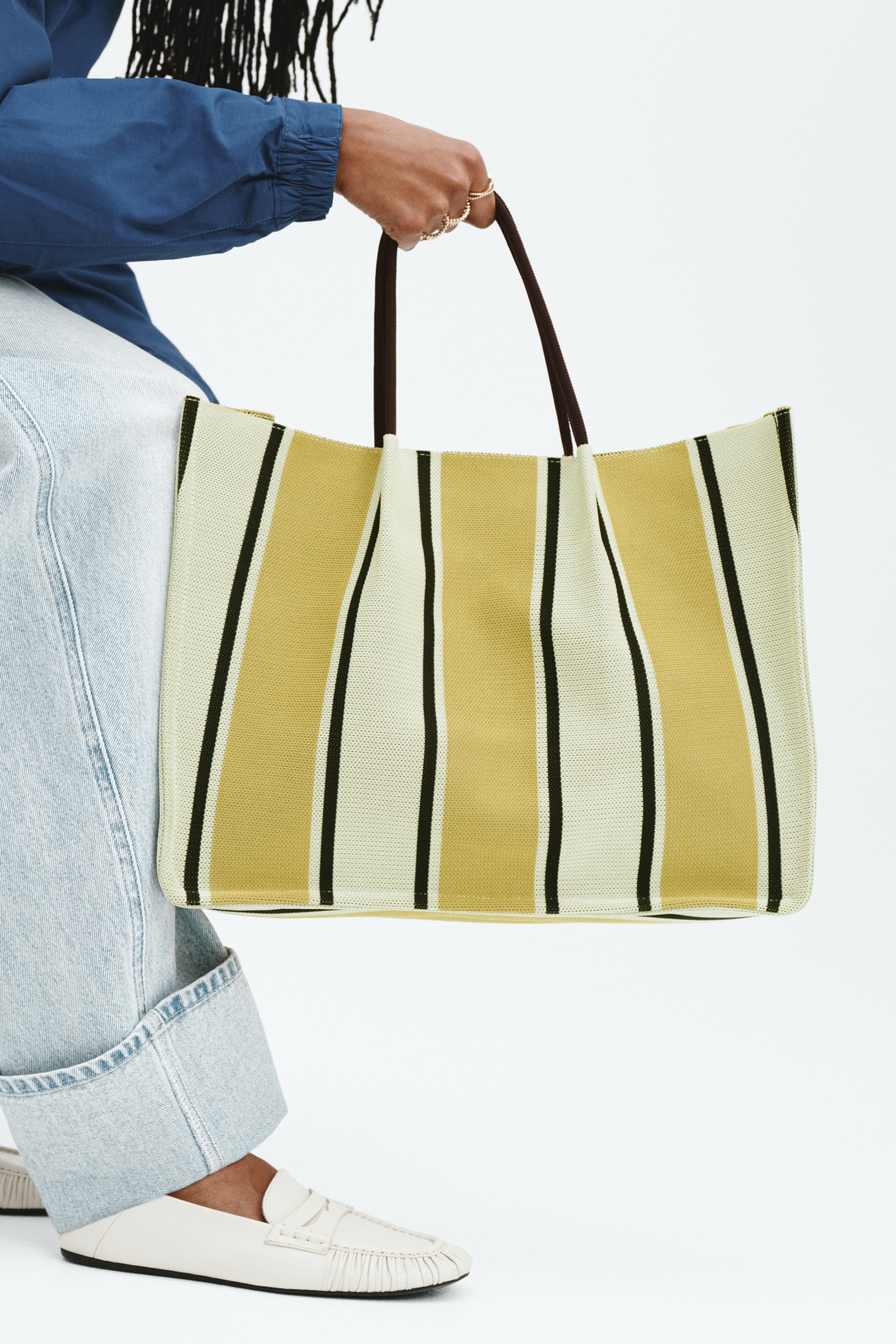Vis større bilde: En solid strikket tote bag med vertikale striper i krem, gul og svart, holdes av modellen som griper de mørkebrune topphåndtakene.