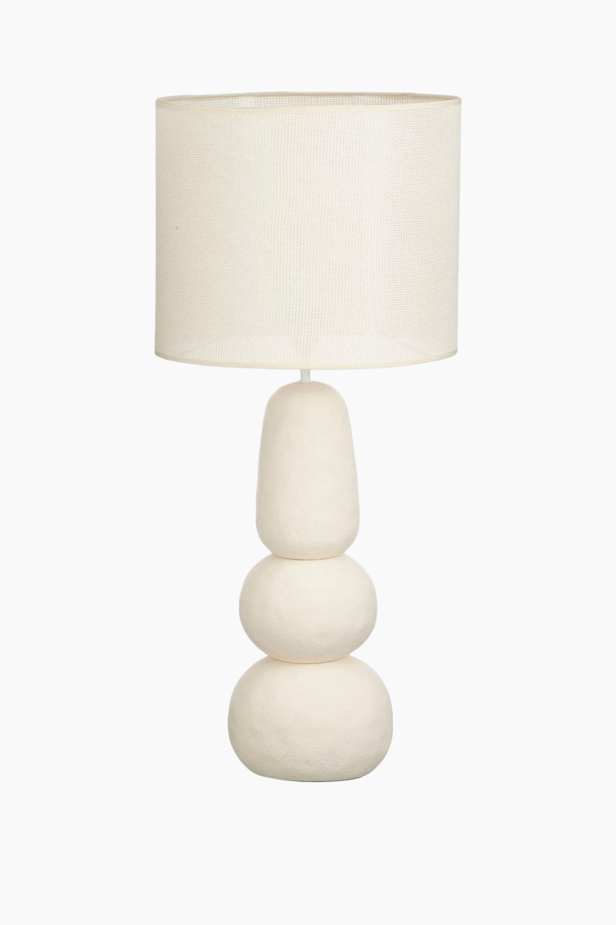 Ver imagen más grande: Se presenta una lámpara de mesa de color crema, que cuenta con una pantalla cilíndrica de tela tejida. Su base está formada por tres formas apiladas, irregulares y de color crema, siendo las dos inferiores esféricas y la superior un óvalo alargado.