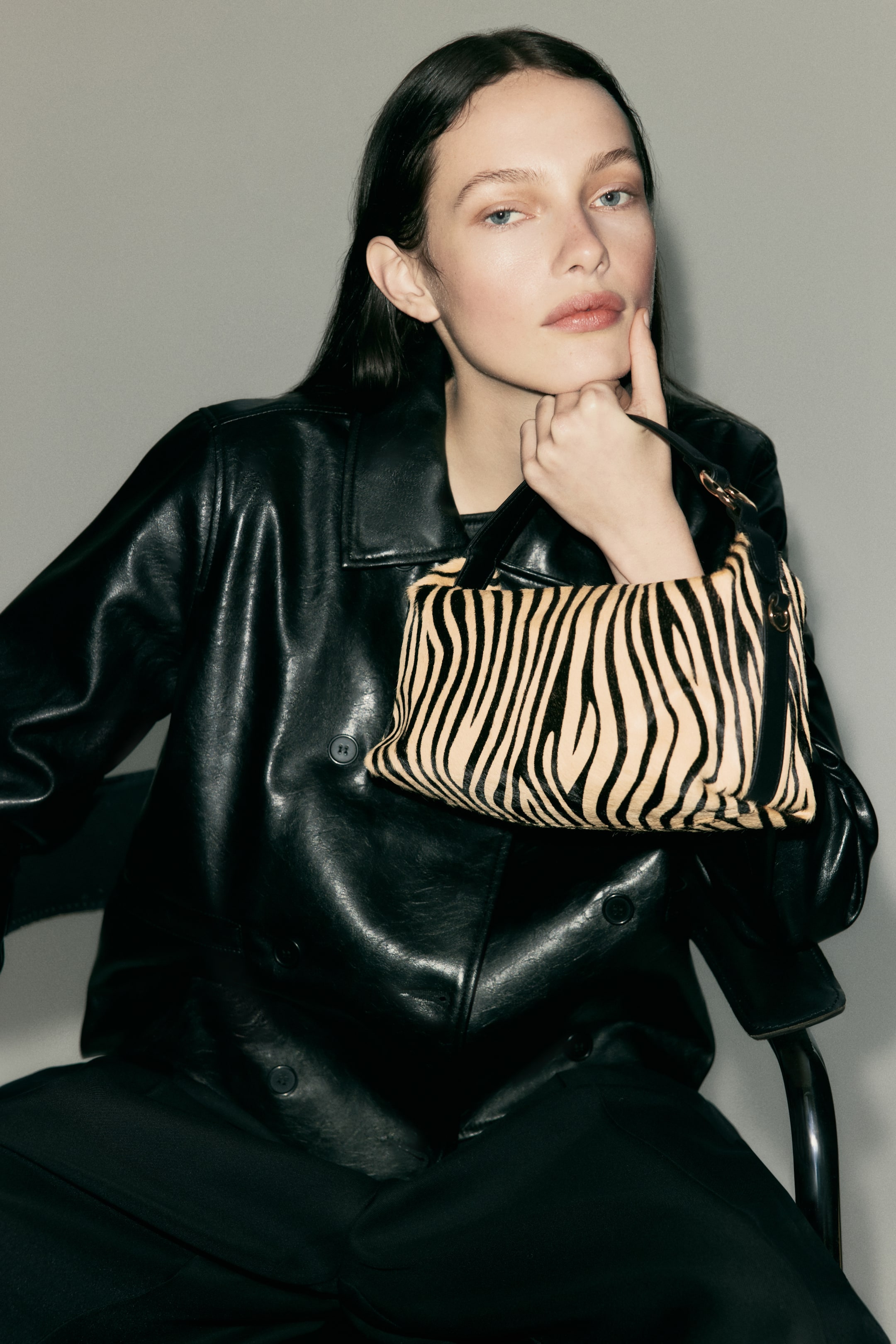 Grotere afbeelding bekijken: Een schoudertas met zebraprint en een geborstelde afwerking wordt vastgehouden door het model. De tas heeft een zwart handvat met een goudkleurige metalen ring aan één kant, en het model houdt de tas vast aan dit handvat, waarbij de tas tegen haar donkere jas rust.