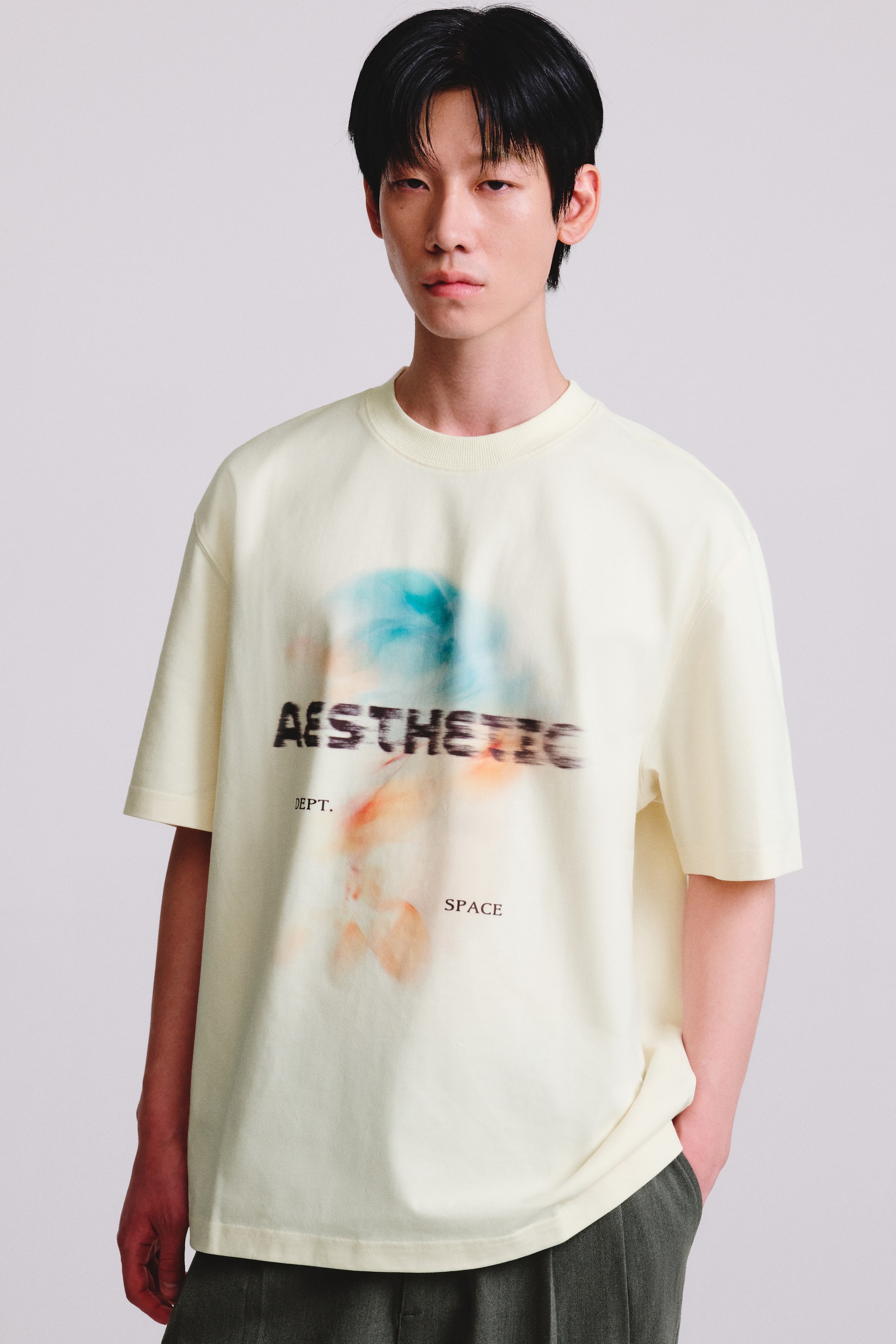 查看大圖: 模特兒身穿一件箱型版型的奶油色 T 恤,採用羅紋圓領、落肩設計,正面印有模糊抽象圖案與文字。這款寬鬆 T 恤搭配深綠色長褲,模特兒左手插在口袋中。