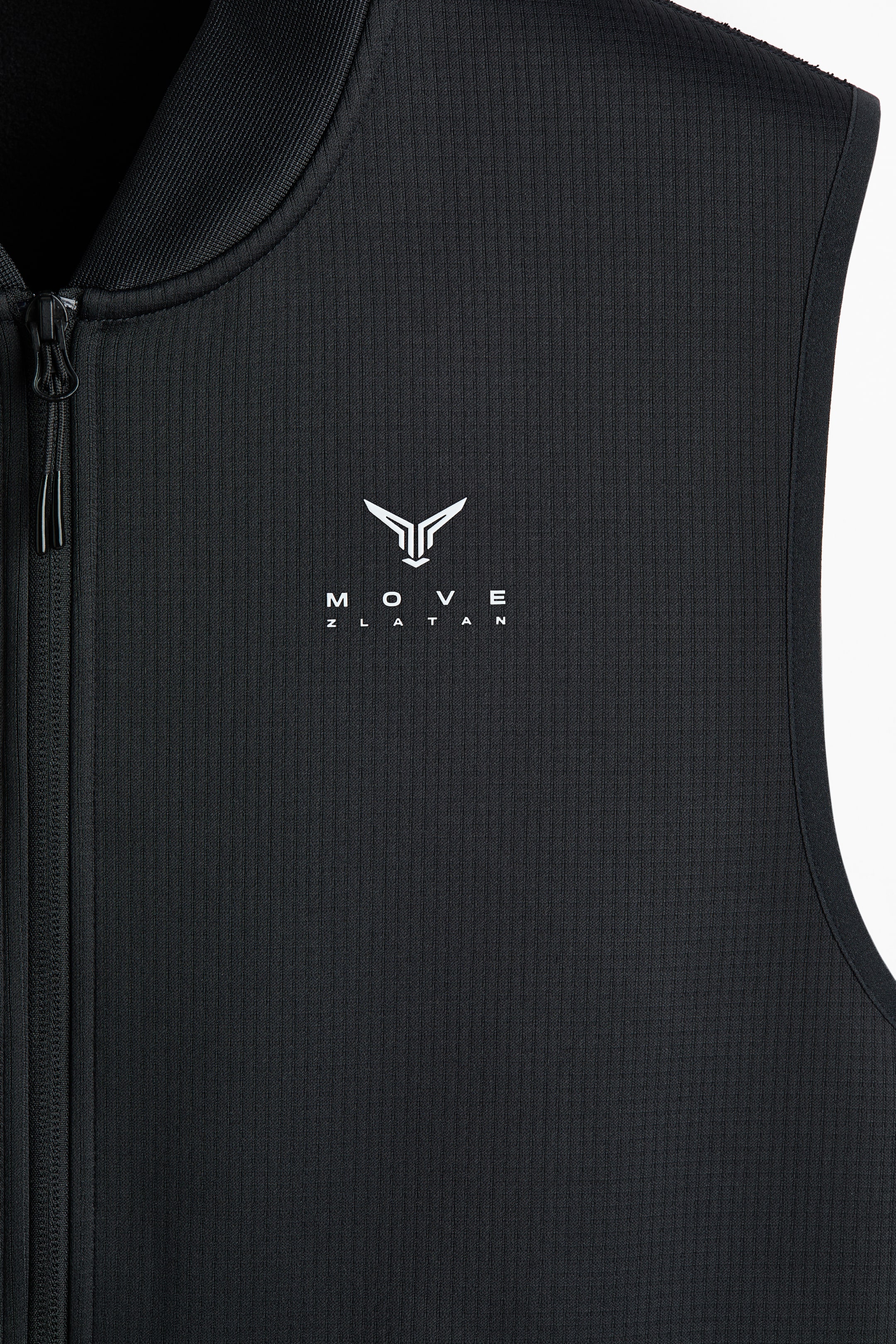 Ampliar la imagen: Un chaleco negro sin mangas está hacia el FRENTE, con una textura acanalada en toda su tela. Muestra un cuello de pie acanalado y un cierre de largo completo que recorre el centro del frente. Un logotipo blanco, que consta de un emblema estilizado y el texto 'MOVE ZLATAN', está posicionado en la parte superior del pecho.