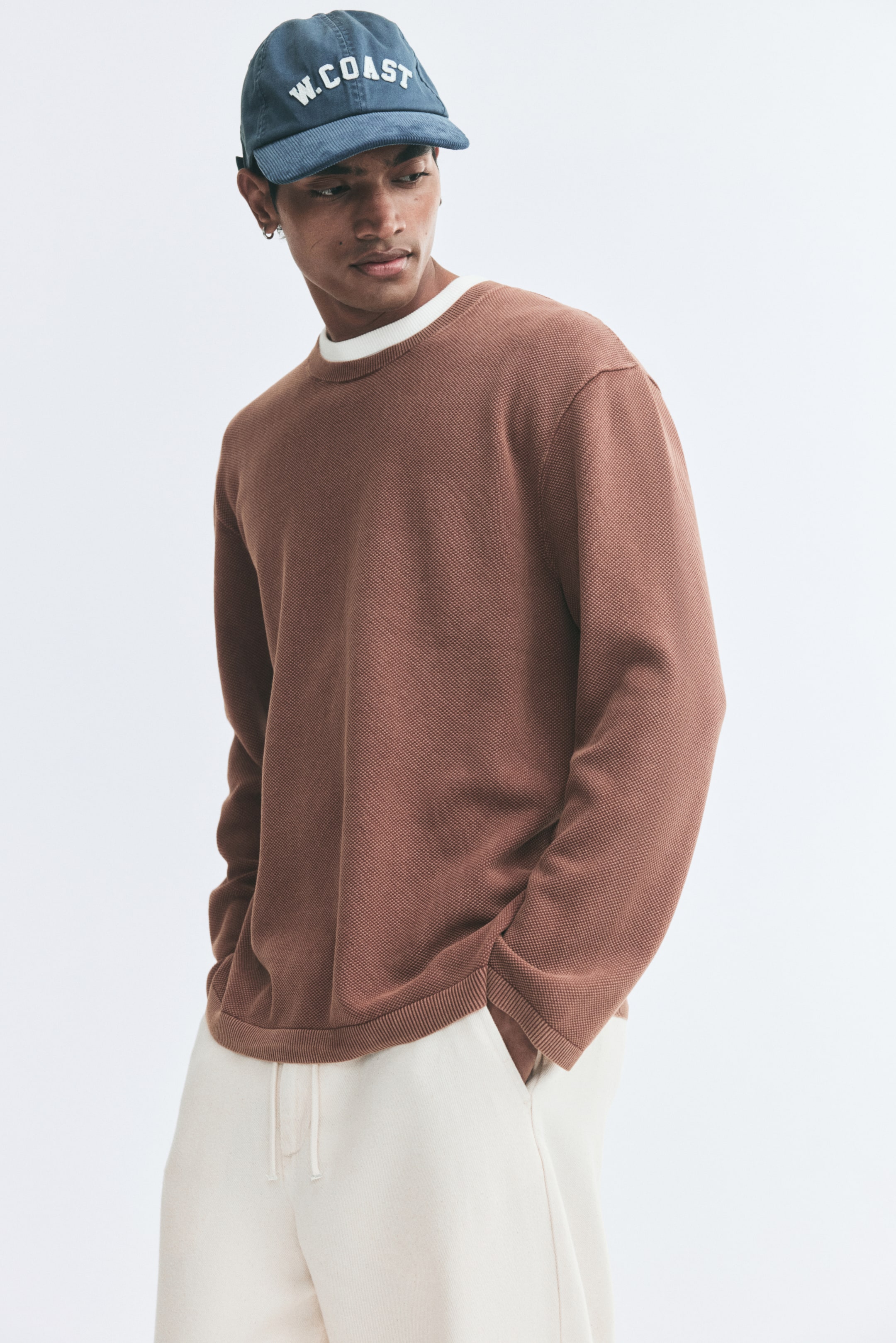 View larger image: A brown textured cotton knit sweater is being worn by the model, featuring a relaxed fit, a round neckline, long sleeves, and ribbed trim. A white T-shirt is visible layered underneath the collar, and the sweater is styled with off-white drawstring trousers and a blue cap.