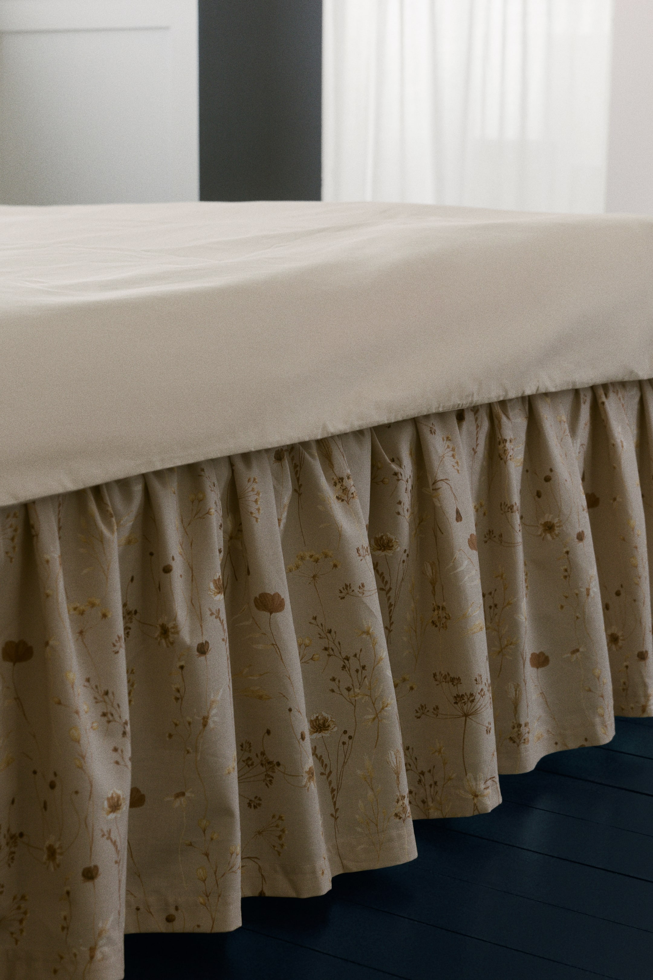 Visa större bild: En volangkant, som syns framifrån, visar ett fint blommönster i nyanser av brunt och guld på en beige bakgrund. Den hänger mjukt och är placerad under ett enkelt ljusfärgat lakan.