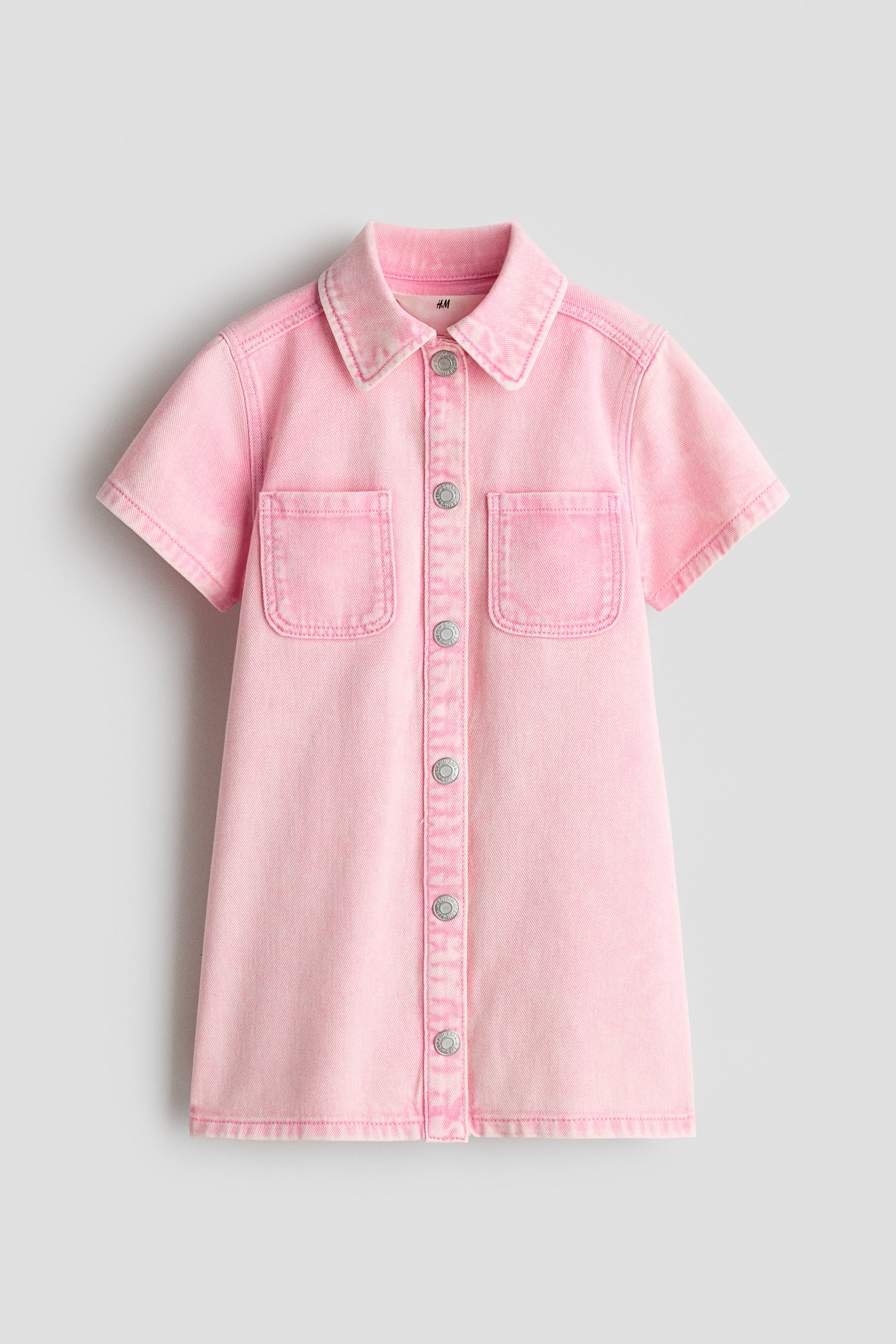 View larger image: A short-sleeved pink cotton denim dress is shown from the front, featuring a classic collar, a button-down front with silver metal buttons, and two open patch pockets on the chest. The dress has a straight, A-line silhouette with a light pink wash, showing subtle fading along the seams and hems.