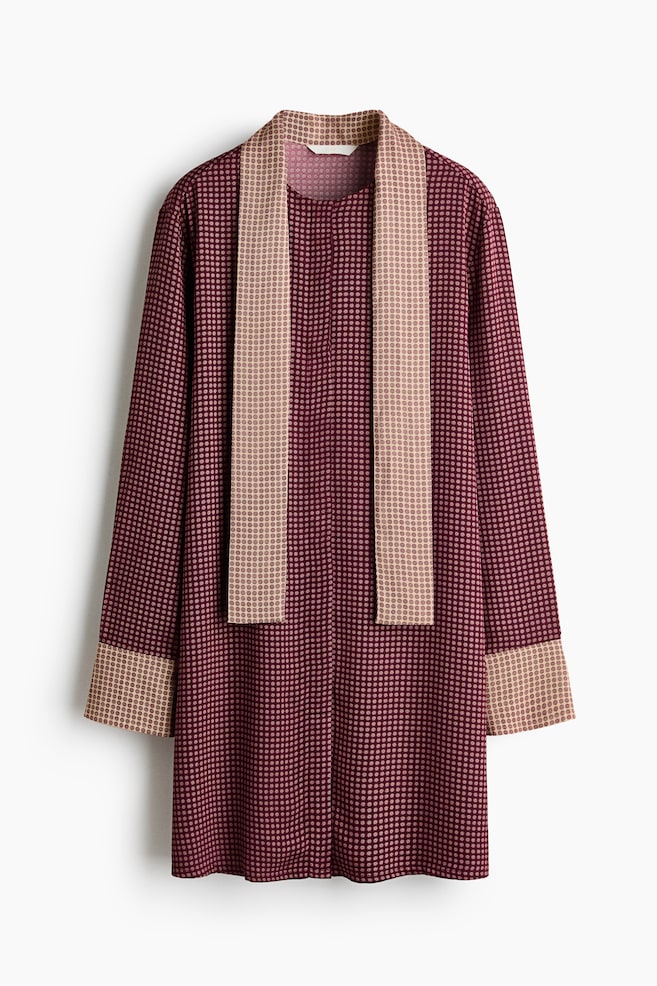 Un vestido rojo oscuro de frente presenta un sutil estampado de puntos y cuenta con un escote redondo con un panel largo en color canela contrastante que cuelga verticalmente por la parte delantera. La prenda también presenta puños anchos; tanto estos puños como el panel muestran un estampado de puntos en color canela a juego. - 1